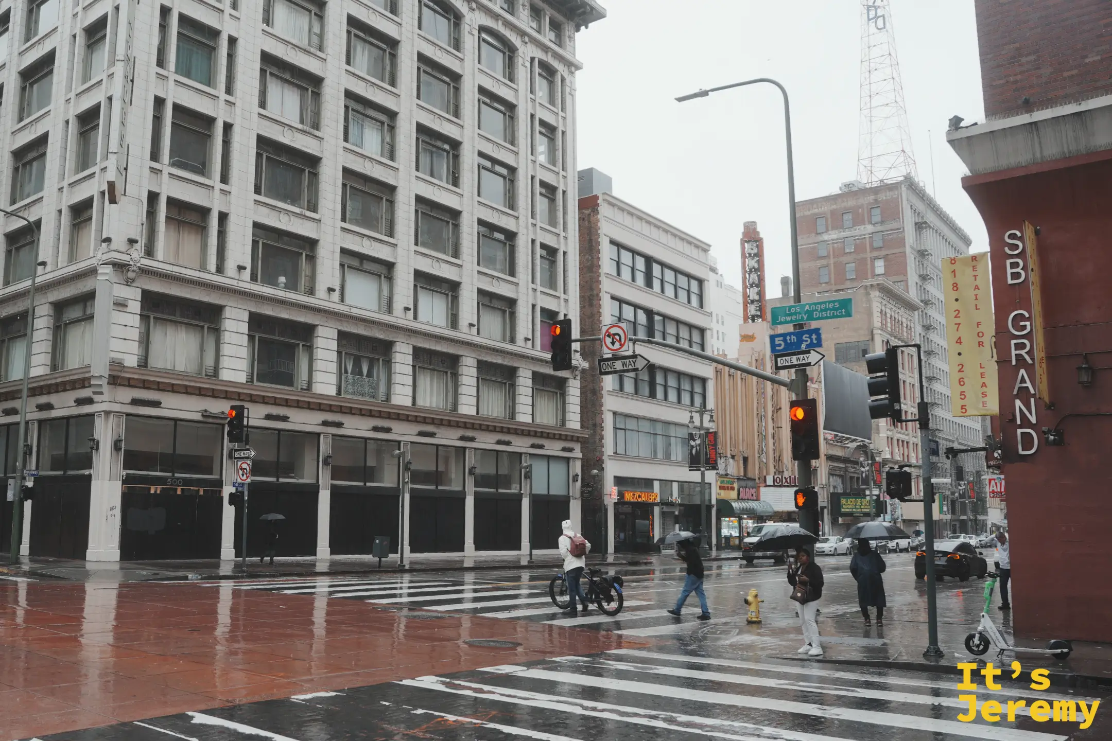 雨中的 DTLA 十字路口，5th St 與 Los Angeles Jewelry District 路牌、SB Grand 紅磚樓與撐傘行人