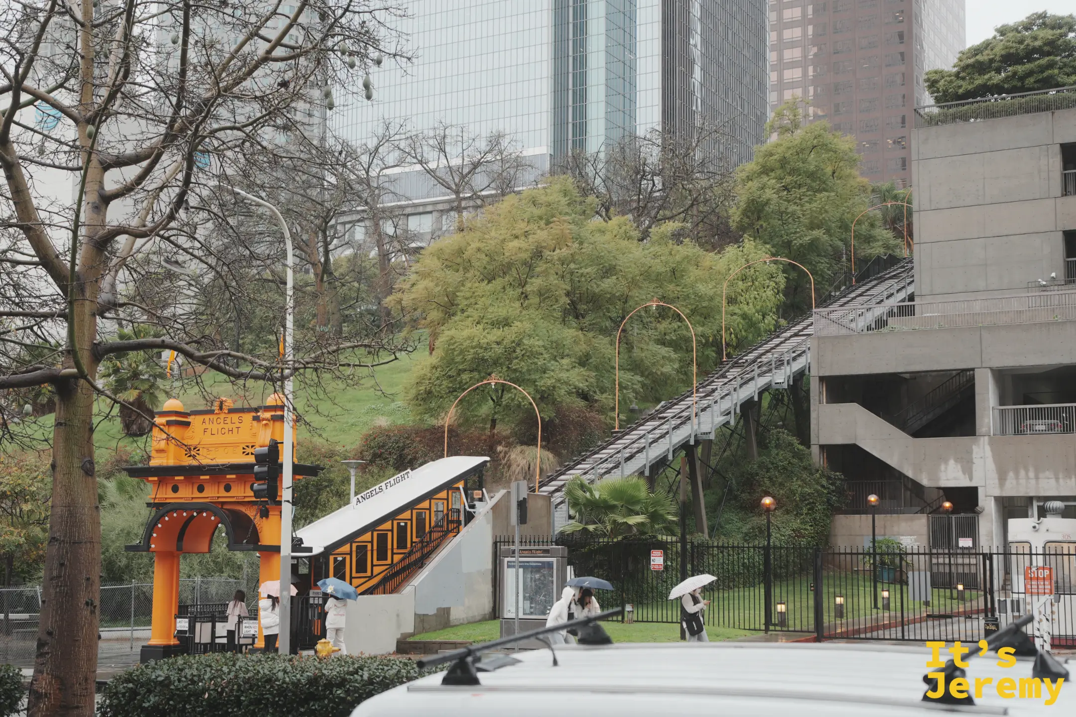 Angels Flight 橘黃色復古纜車與黃色拱門站體，陡坡軌道通往山上，雨天撐傘的旅人
