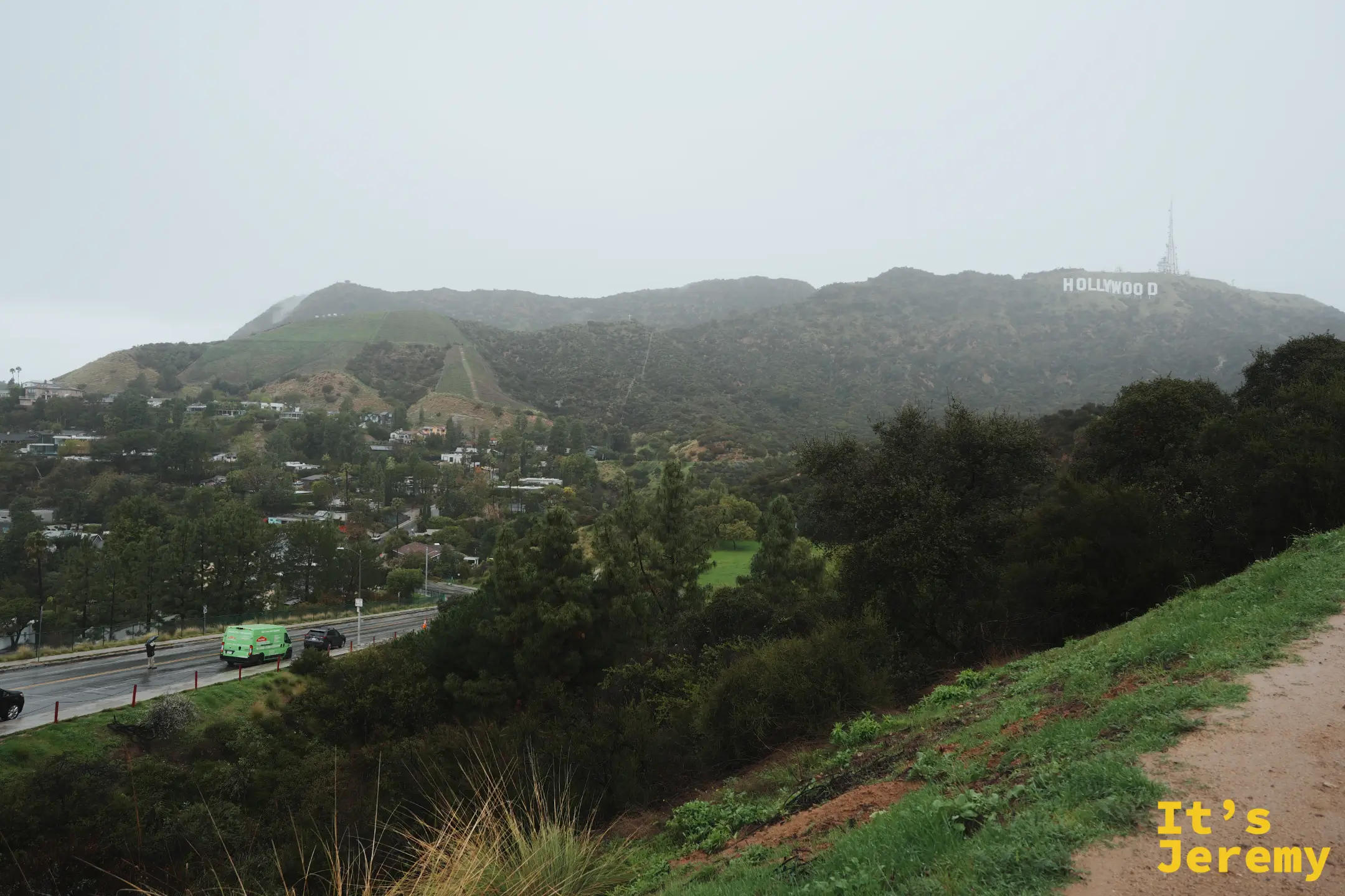 隔著山谷遠眺好萊塢標誌全景，前景是山坡步道與住宅區