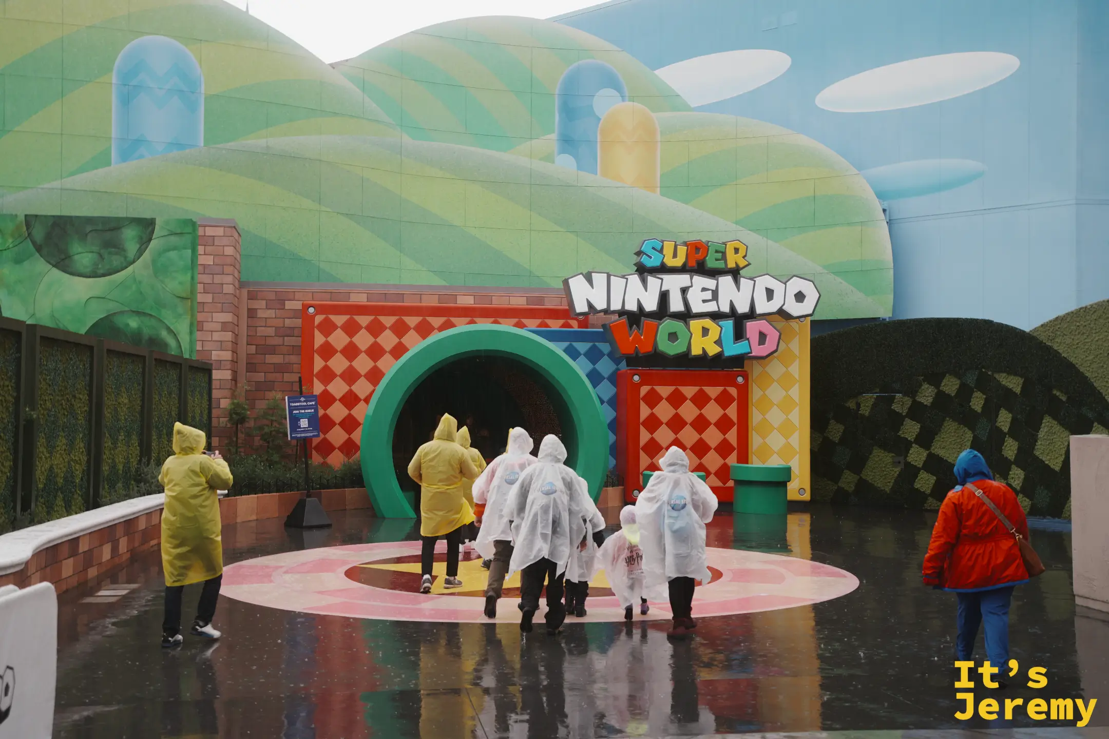 Super Nintendo World 入口的綠色水管隧道與招牌，遊客穿雨衣走入