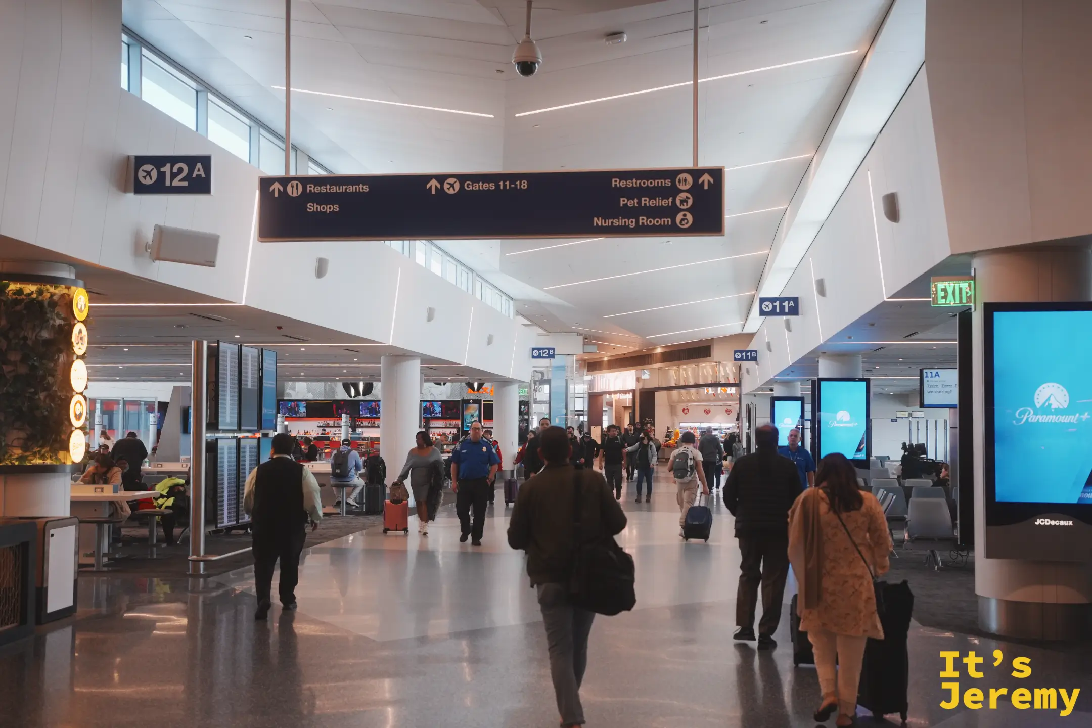 LAX 候機廳走道，頭頂懸掛 Restaurants、Shops、Gates 11-18、Restrooms 的指標，旅客拖著行李往登機門走