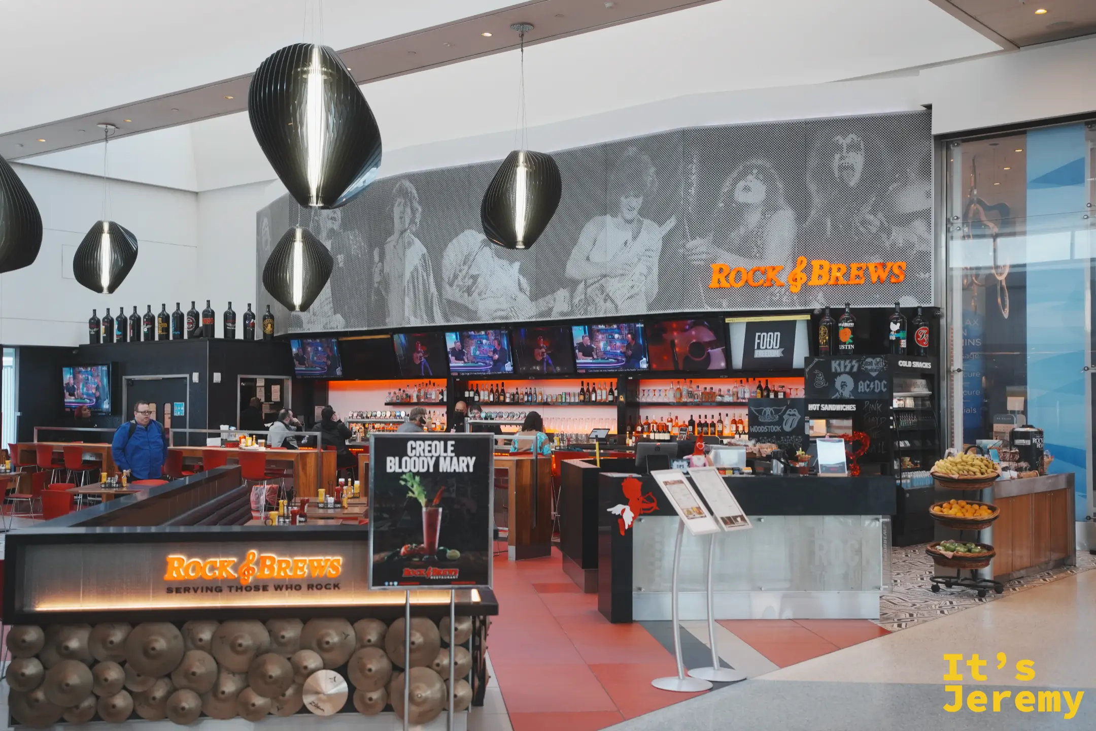 LAX 的 Rock & Brews 搖滾主題餐酒吧，牆上是黑白搖滾巨星壁畫、吧台以鈸片裝飾，立牌寫著 Creole Bloody Mary