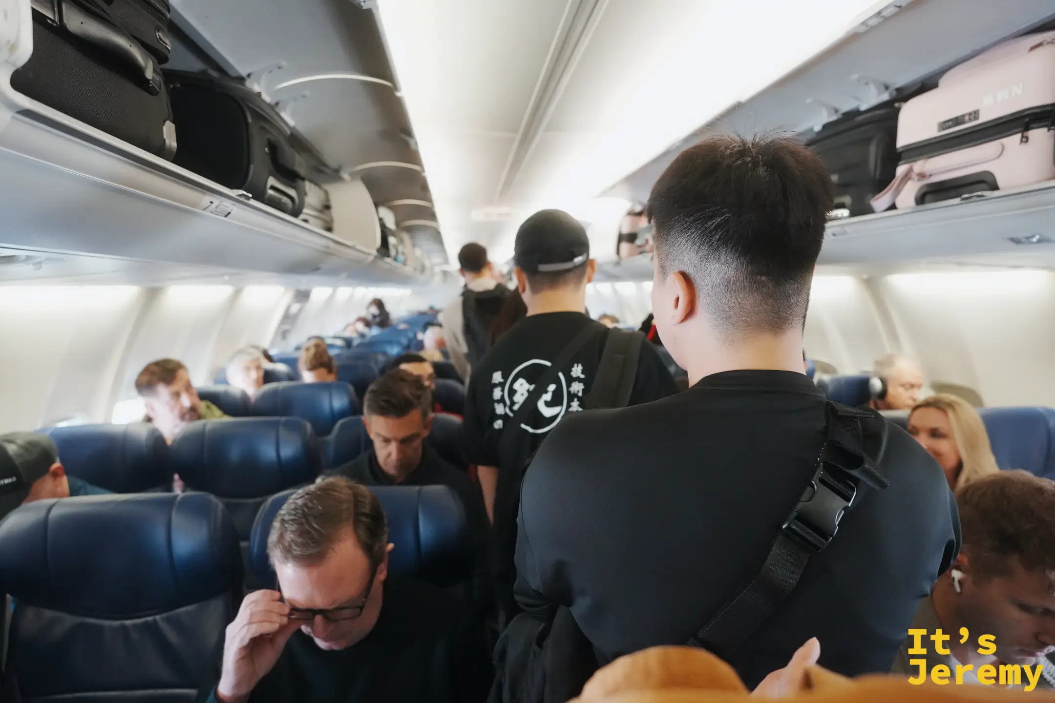 南西航空機艙走道，旅客拖著背包與行李陸續往機艙後段移動，頭頂置物櫃塞滿手提行李