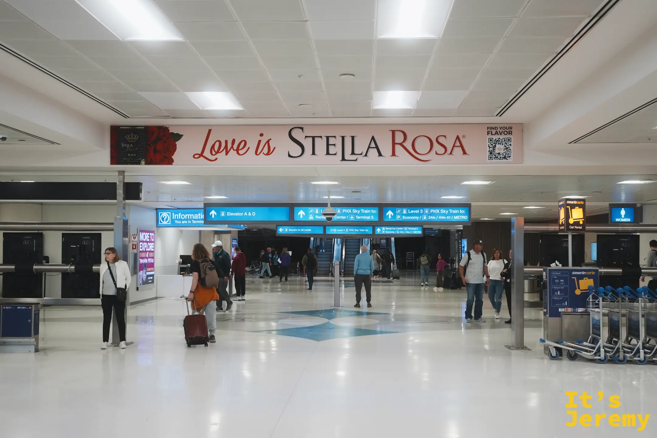 鳳凰城天港機場航廈內，Love is Stella Rosa 廣告橫幅下方是 PHX Sky Train 接駁指標與成排的 SmarteCarte 行李推車