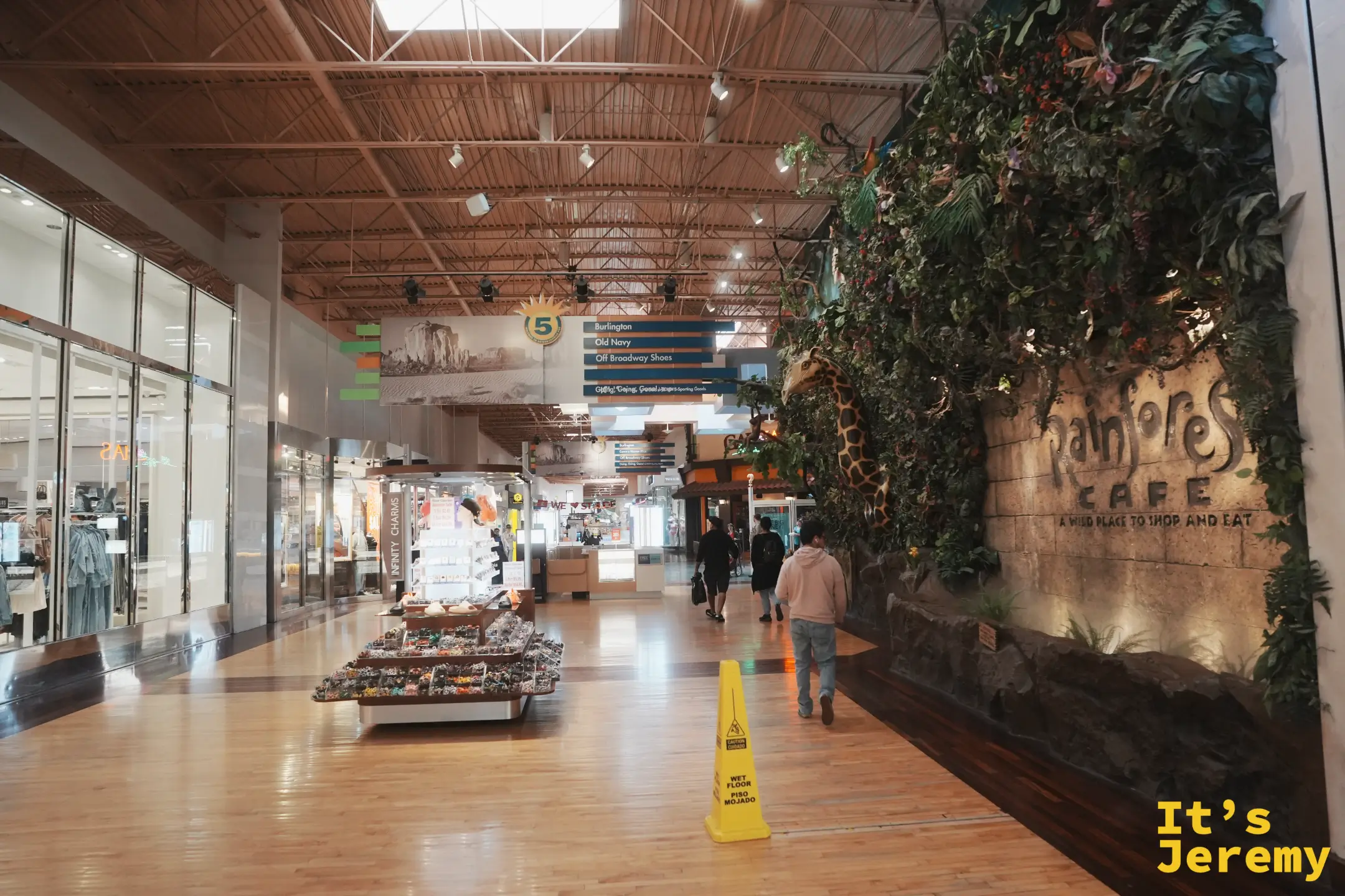 Arizona Mills 商場主走道，右側是 Rainforest Cafe 的雨林綠牆與探頭的長頸鹿，頭頂懸掛 Burlington、Old Navy 的指標