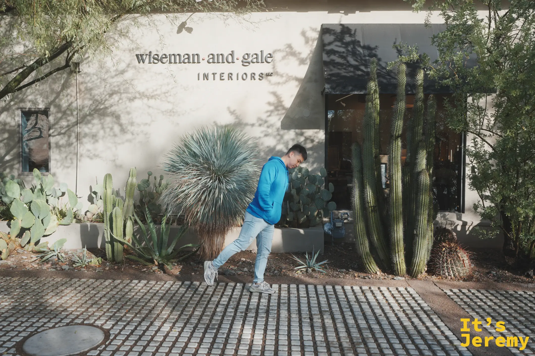 Jeremy 穿藍色帽 T 走過 wiseman and gale interiors 門前的沙漠植栽，柱狀仙人掌與龍舌蘭在陽光下投下樹影