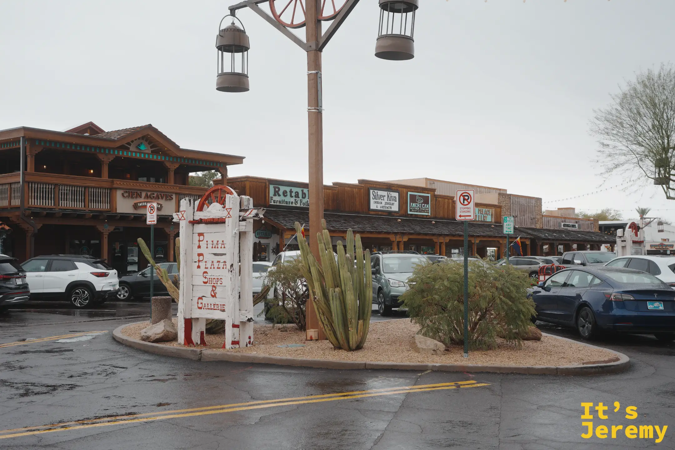 雨中的 Pima Plaza Shops & Galleries 白色木招牌與馬車輪路燈，後方是 Cien Agaves 等木造西部店面