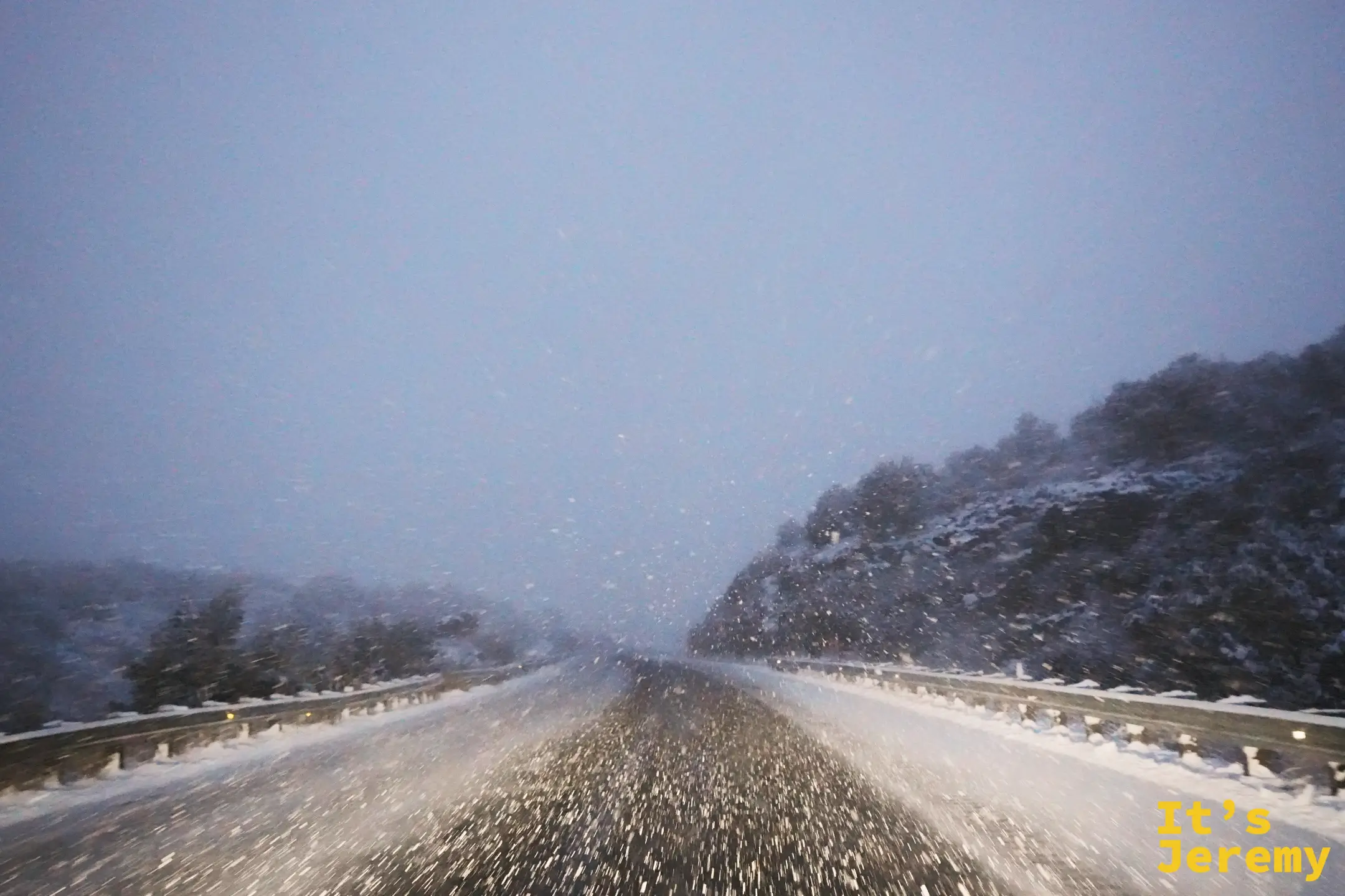 高速公路上的暴雪，路面積著雪水、雪花斜打在擋風玻璃上，右側山坡與護欄邊都積了雪，能見度很低