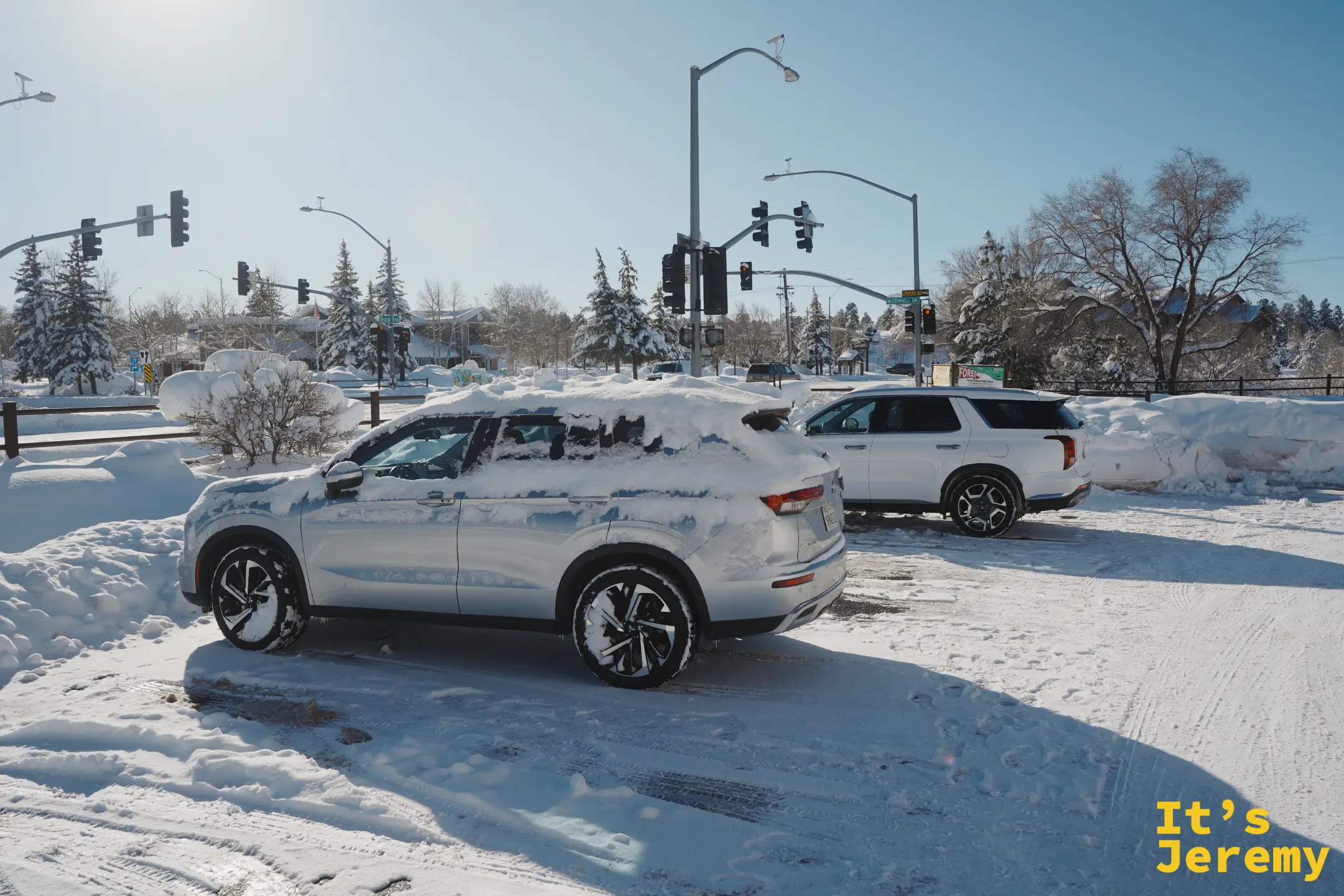 停車場裡一台被雪覆蓋的銀色休旅車停在雪地裡，藍天下四周都是積雪的松樹與路口號誌