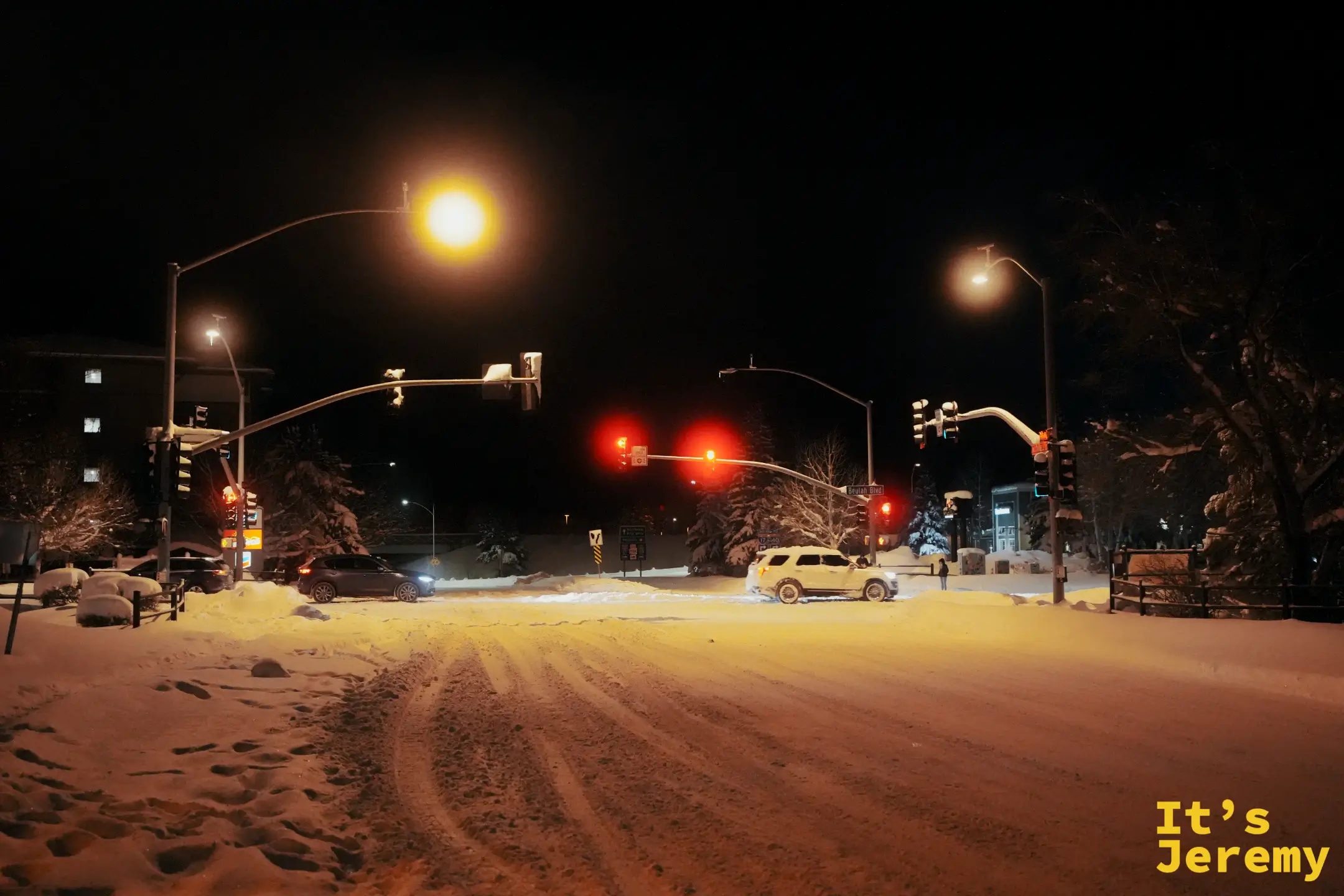 Flagstaff 雪夜的十字路口，路口積著厚雪、路面壓出車轍，號誌燈與街燈的暖光把雪地染成橘黃色