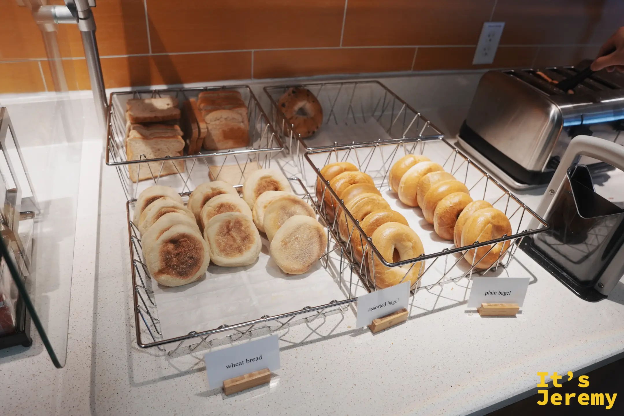 旅館早餐吧的麵包區，鐵籃裡分別放著 wheat bread、英式馬芬、各式貝果，旁邊放著烤麵包機