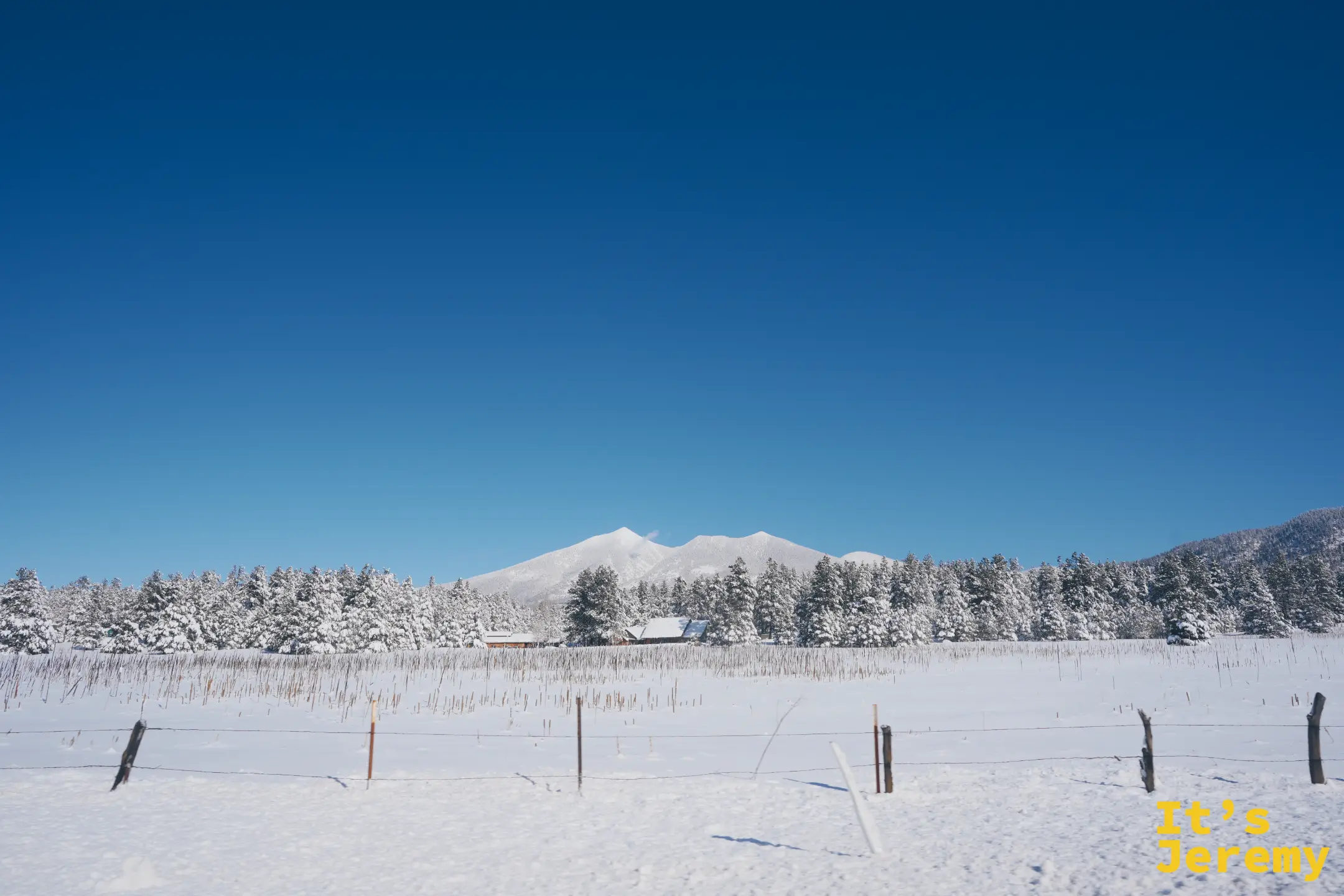 開闊的雪原盡頭，聖弗朗西斯科山峰頂著白雪橫在藍天下的地平線，前景是雪地與圍欄
