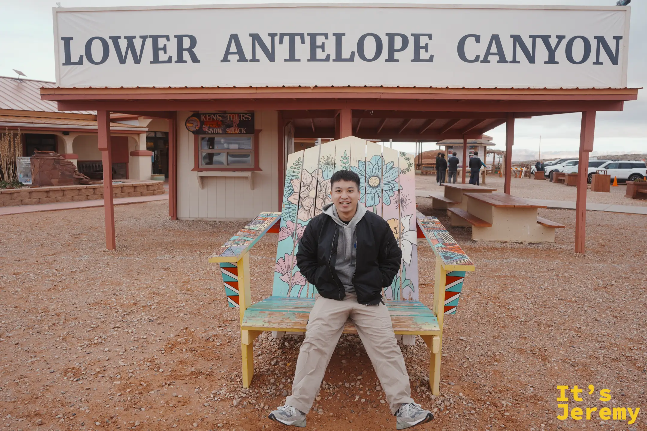 Jeremy 坐在接待區彩繪的大木椅上，身後就是 LOWER ANTELOPE CANYON 的白色招牌