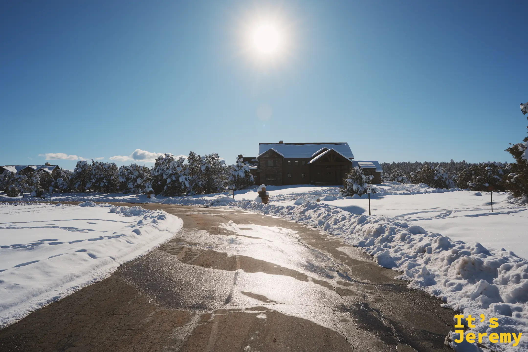 一棟大型石造木屋主建築藏在掛滿雪的樹後，湛藍天空中掛著刺眼的太陽，車道被鏟出一條融雪的路