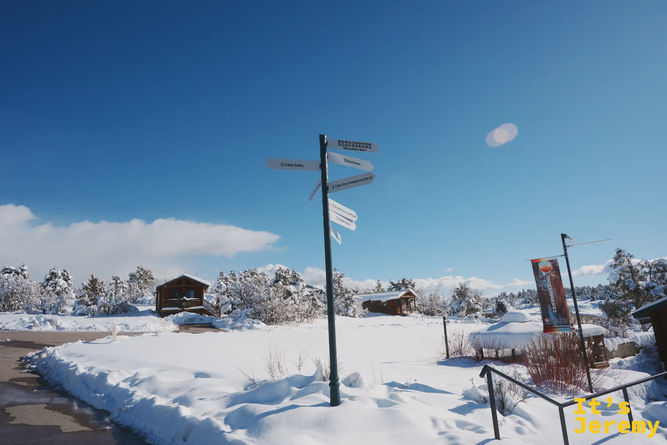 一根掛滿方向箭頭的指標柱立在雪地裡，寫著 Cabin Suites、Restrooms、Recreation Barn、Zion Crest Campground，旁邊有 CANYONEERING 旗幟與雪覆的小木屋