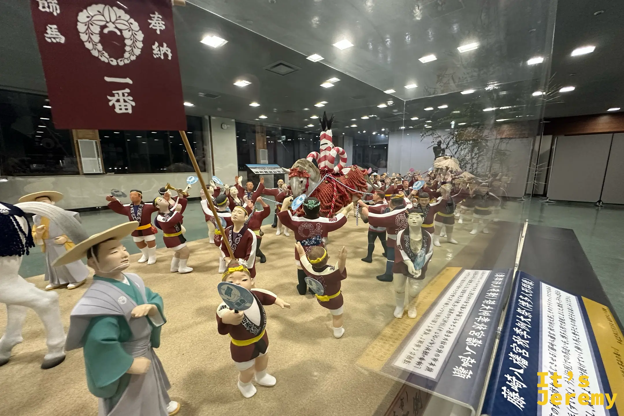 玻璃櫃重現藤崎八旛宮秋季例大祭的『飾馬奉納』，紅色旗幟寫著『奉納 飾馬 一番』，人偶群像簇擁著披紅飾的白馬