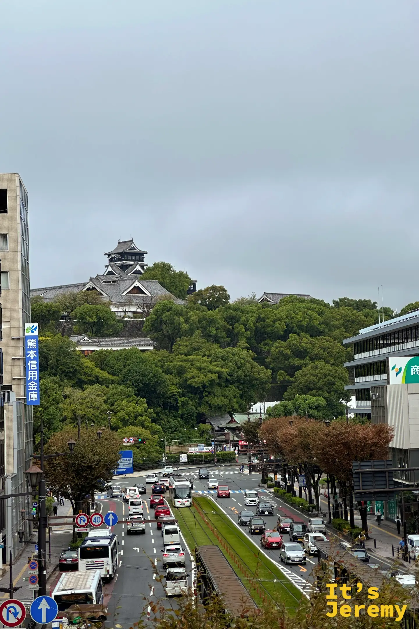 白天陰天下的熊本城天守閣，被大片綠樹環繞，左側是熊本信用金庫的直式招牌