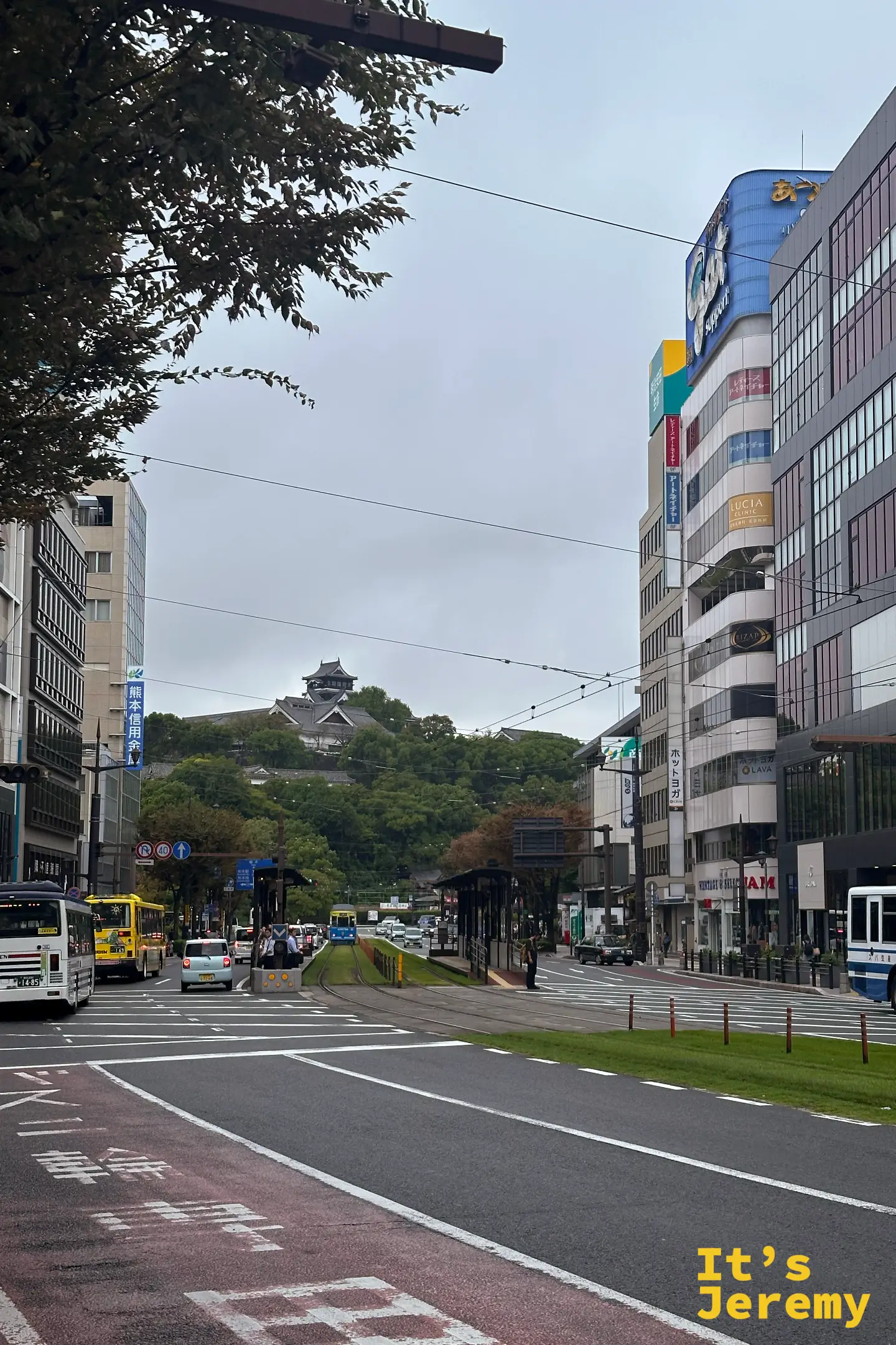 白天的熊本市電大馬路，路面電車與多輛公車在城下交會，遠處是綠意中的熊本城天守閣