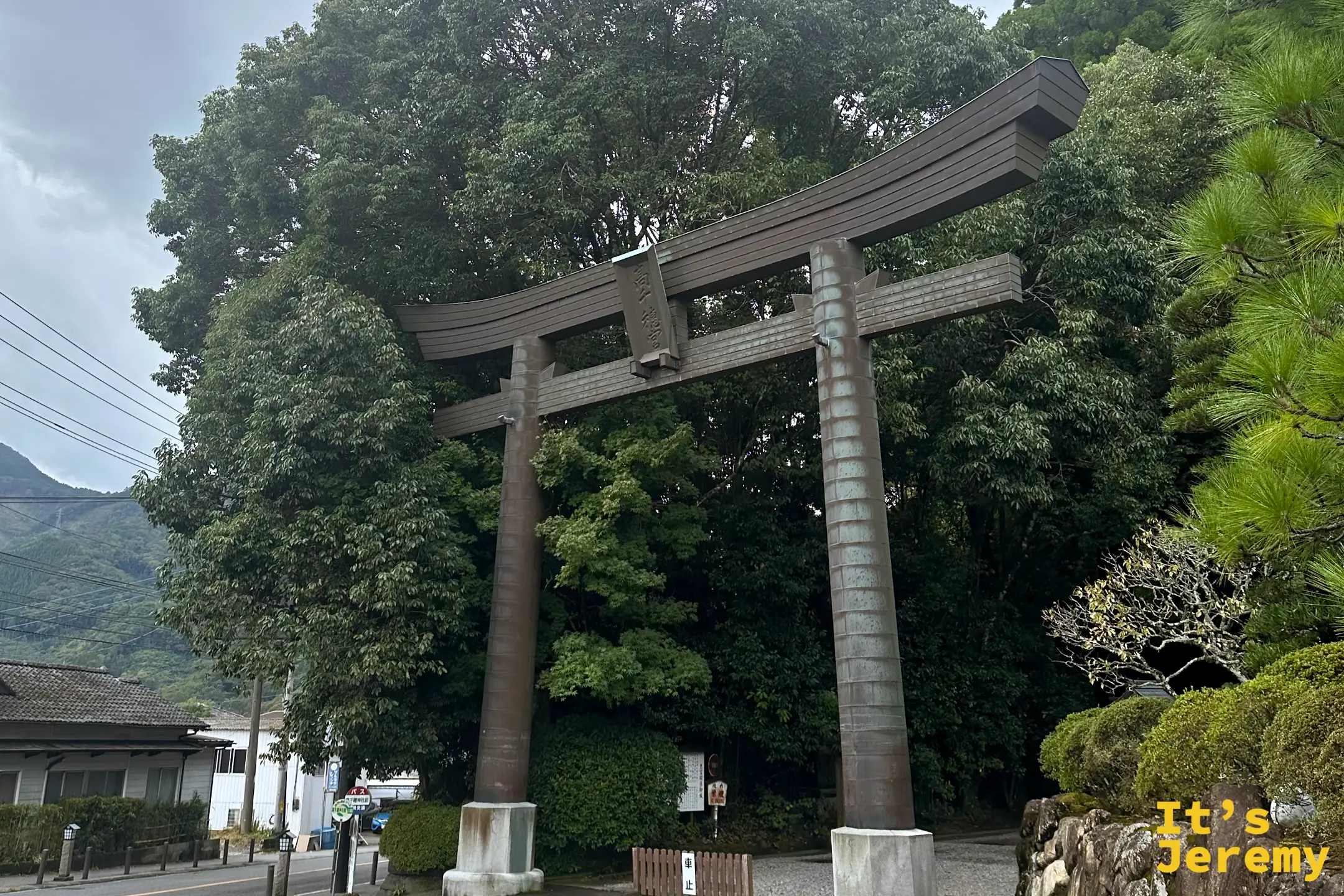 高千穂神社門前的青銅色大鳥居，鳥居後方是連綿起伏、被雲霧輕掩的九州山巒
