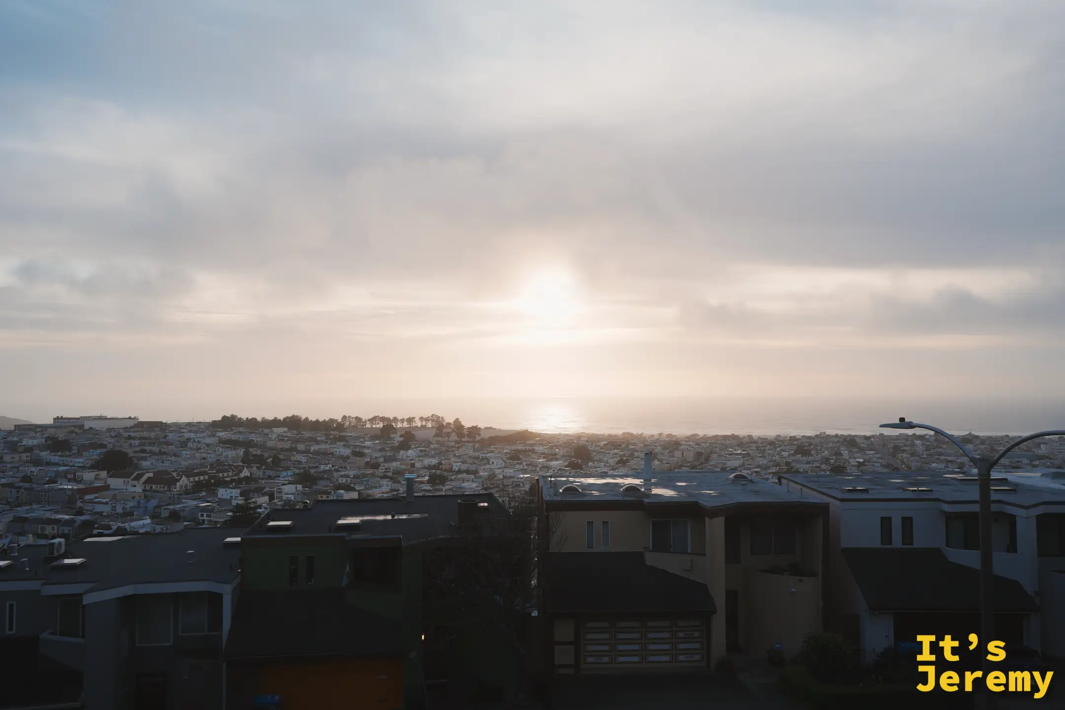 夕陽落在海平面上，光在海面拉出一道亮線，前景是層層疊疊的住宅屋頂