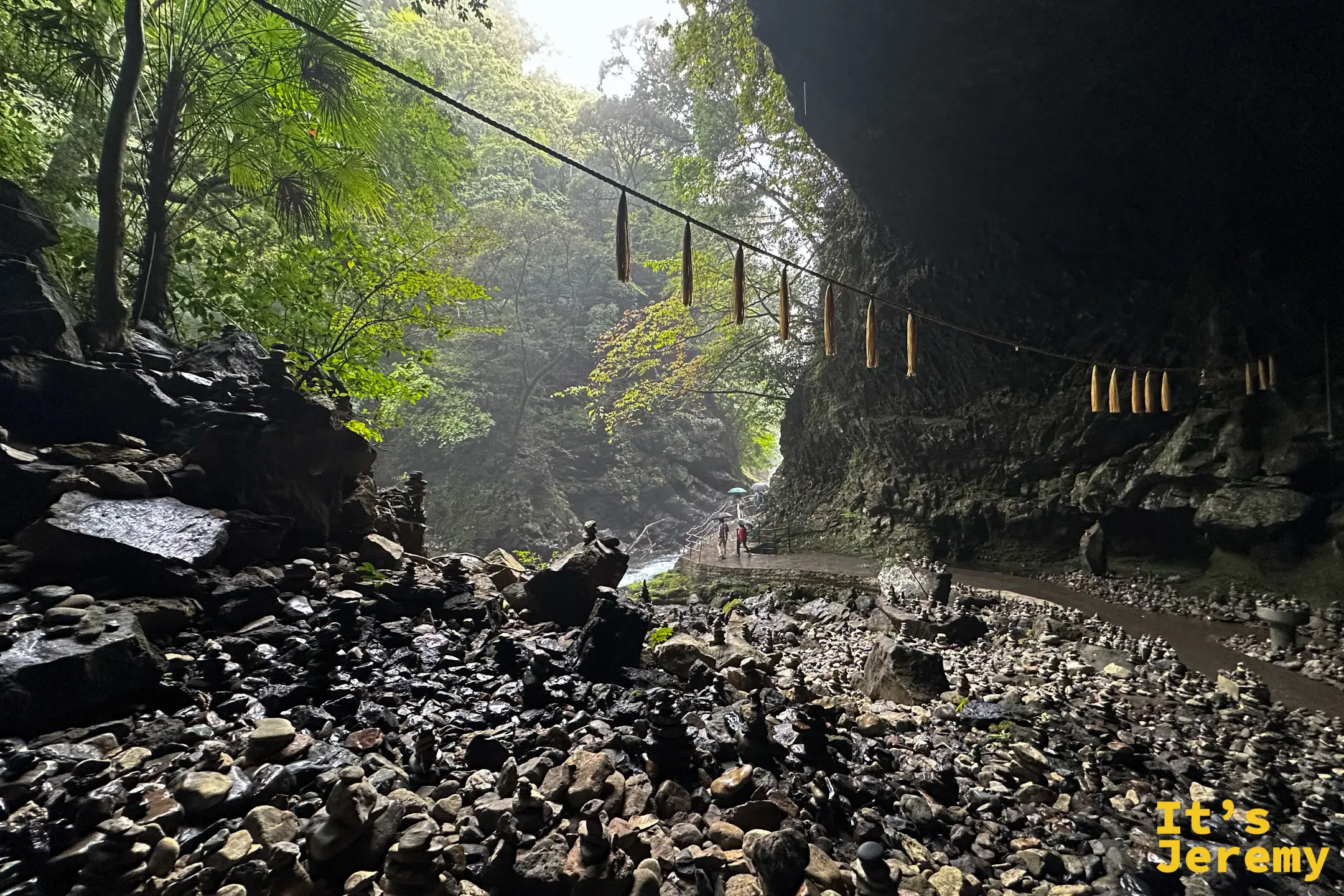 天安河原洞窟內滿地的石堆，注連繩橫過上方，遠處遊客撐傘走過
