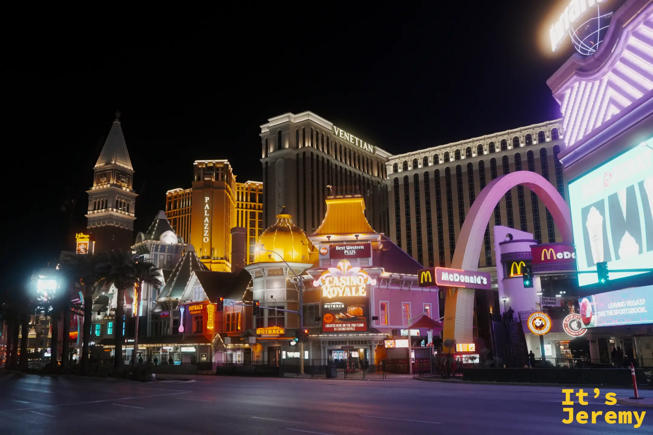 大道另一側的威尼斯人與 Palazzo 外牆打上暖金光，前方是 Casino Royale 與拱形的 McDonald