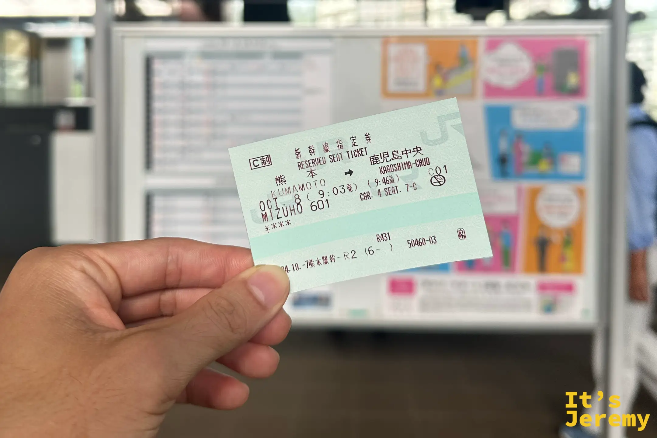 手持一張新幹線指定席車票，起訖是熊本到鹿兒島中央，車種是 Mizuho（みずほ）號，背景是月台的時刻表看板