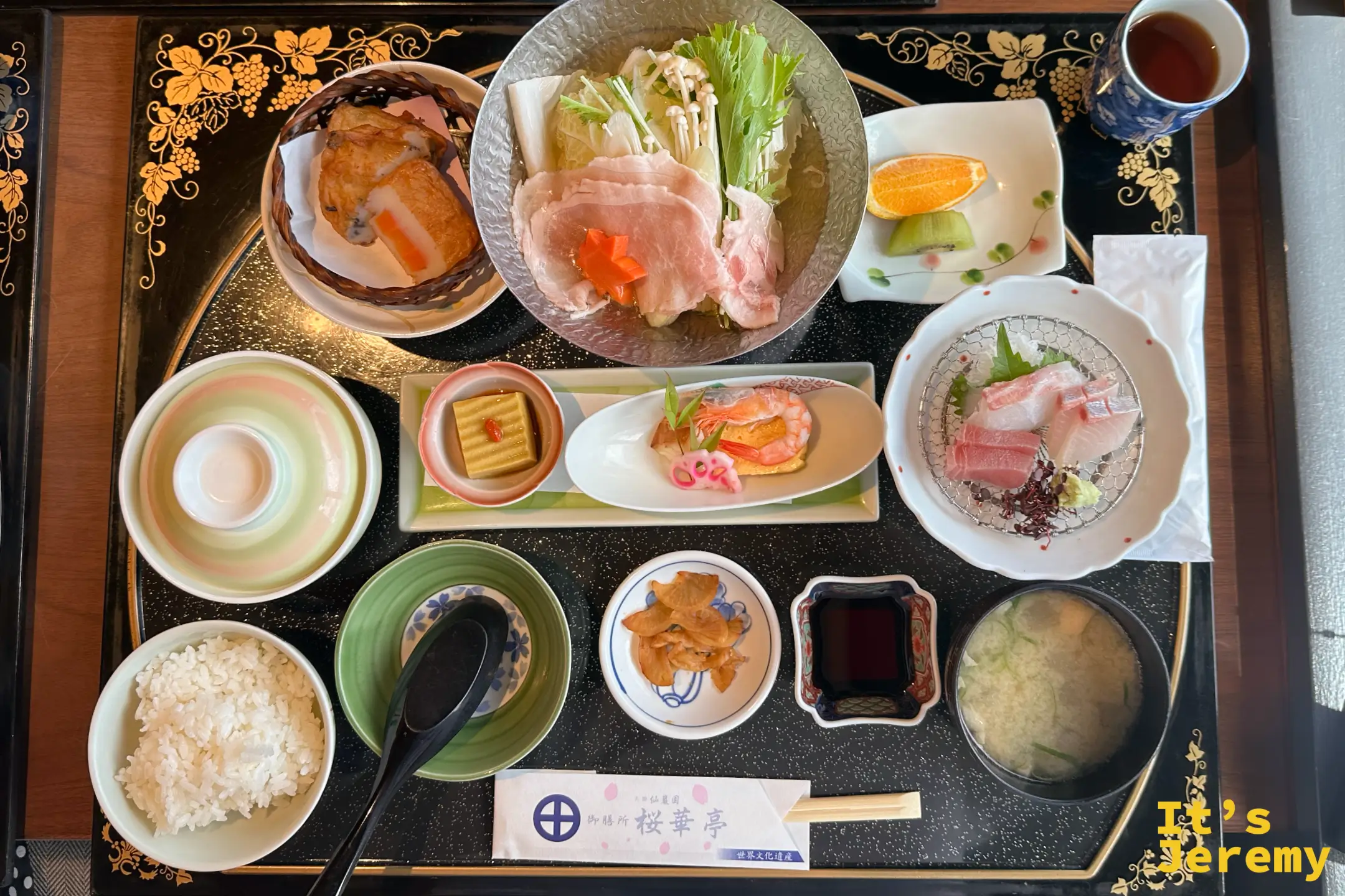 俯拍薩摩郷土料理御膳，黑金漆盤上有黑豚涮涮鍋、生魚片、炸物、白飯、味噌湯與多樣小缽，筷套印著御膳所桜華亭世界文化遺産字樣