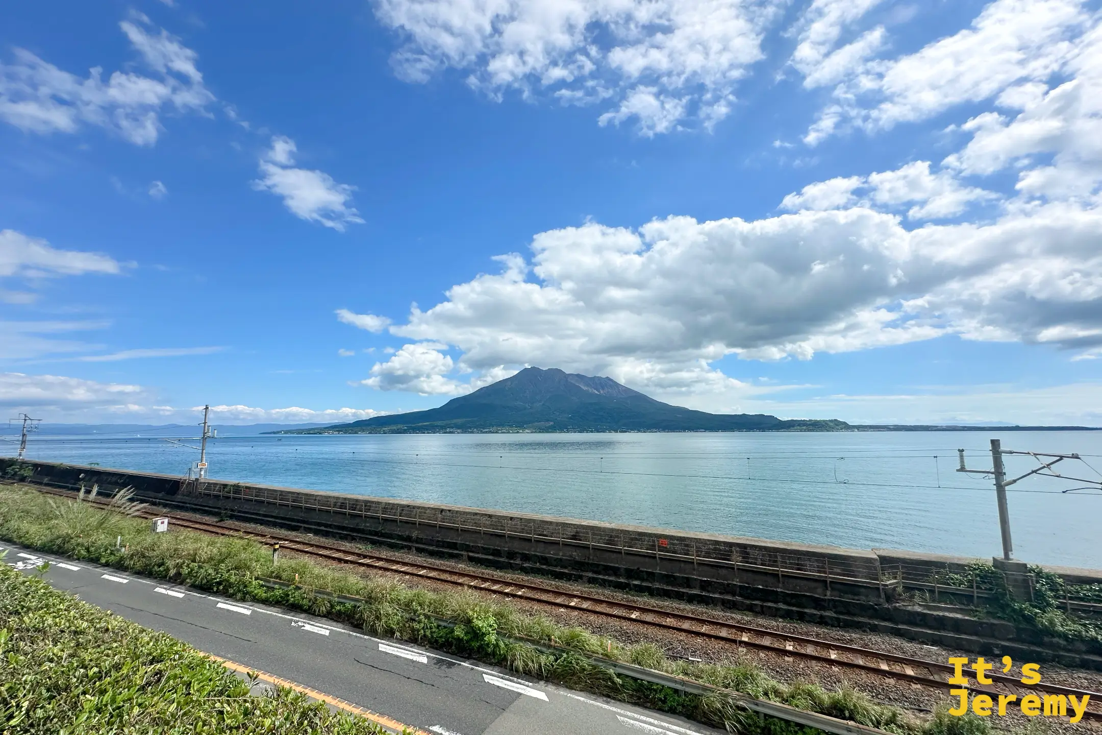 從仙巌園海側望向櫻島，前景是沿海的 JR 鐵道，火山橫亙錦江灣對岸