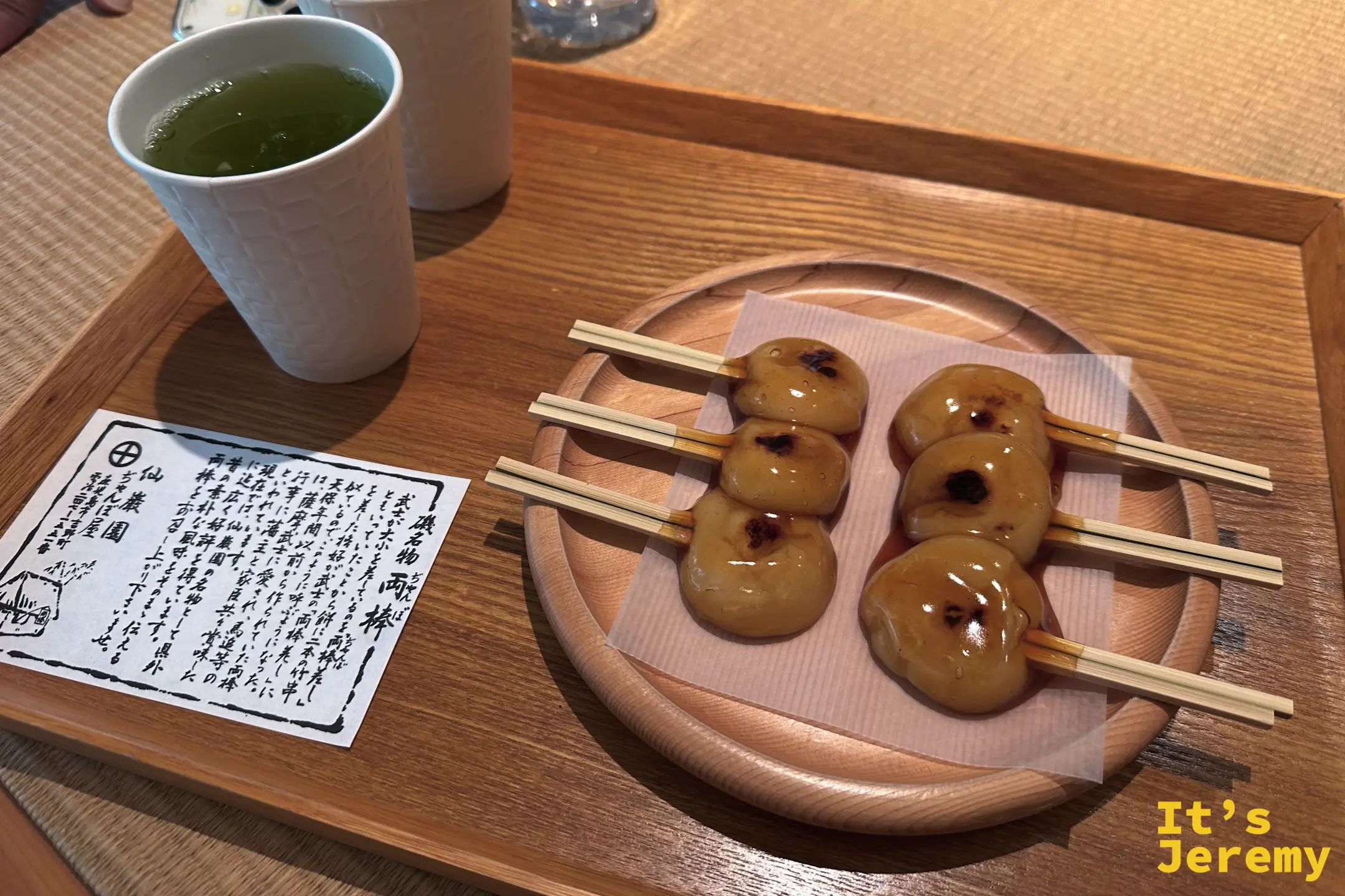 木盤上擺著両棒餅，軟糯的小麻糬每份插著兩根竹籤、裹著焦香醬油醬汁，一旁是一杯綠茶與手寫的名物說明卡