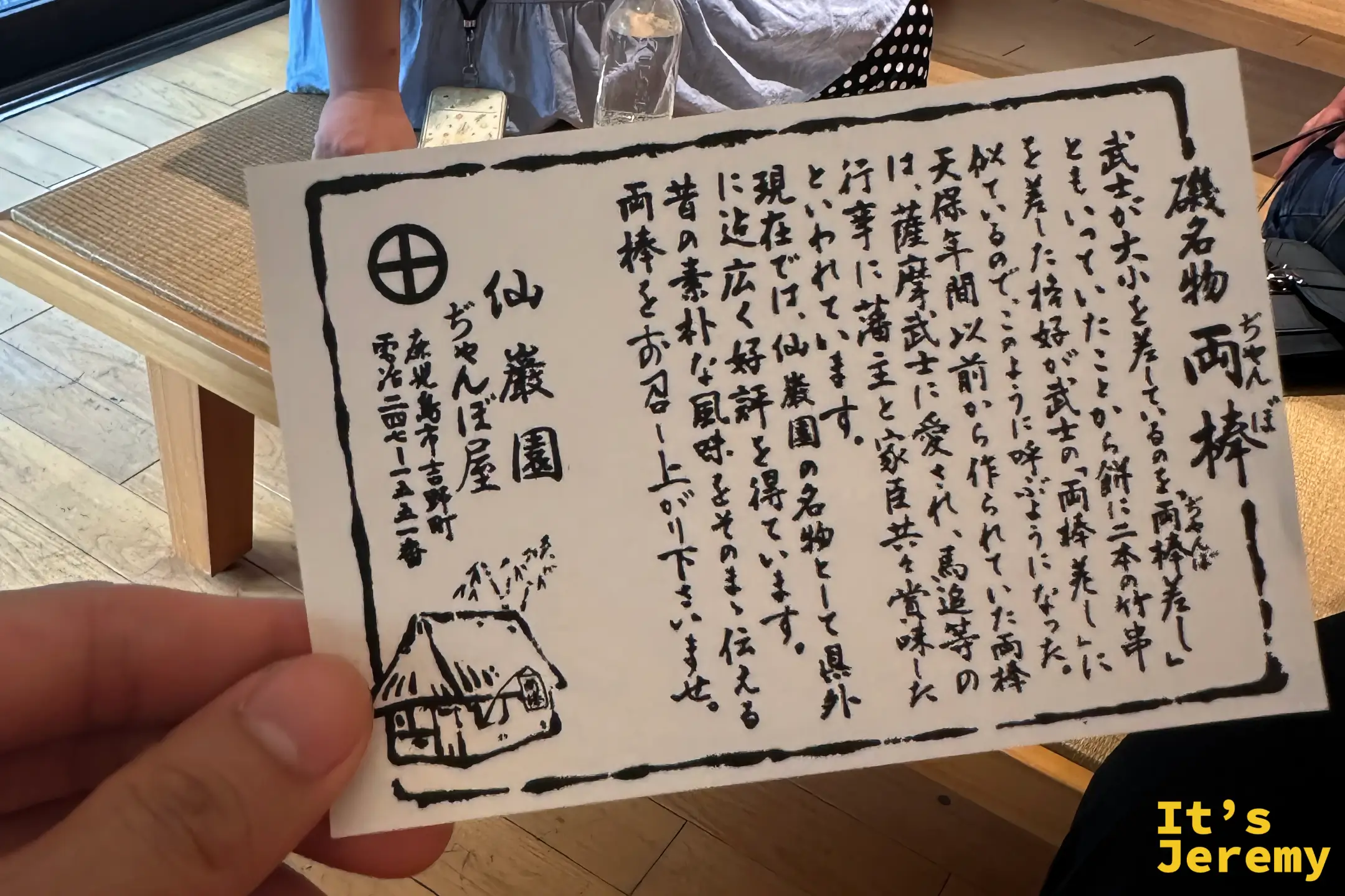 手持ぢゃんぼ屋的説明卡，上面以毛筆字介紹「磯名物 両棒」的由來與典故