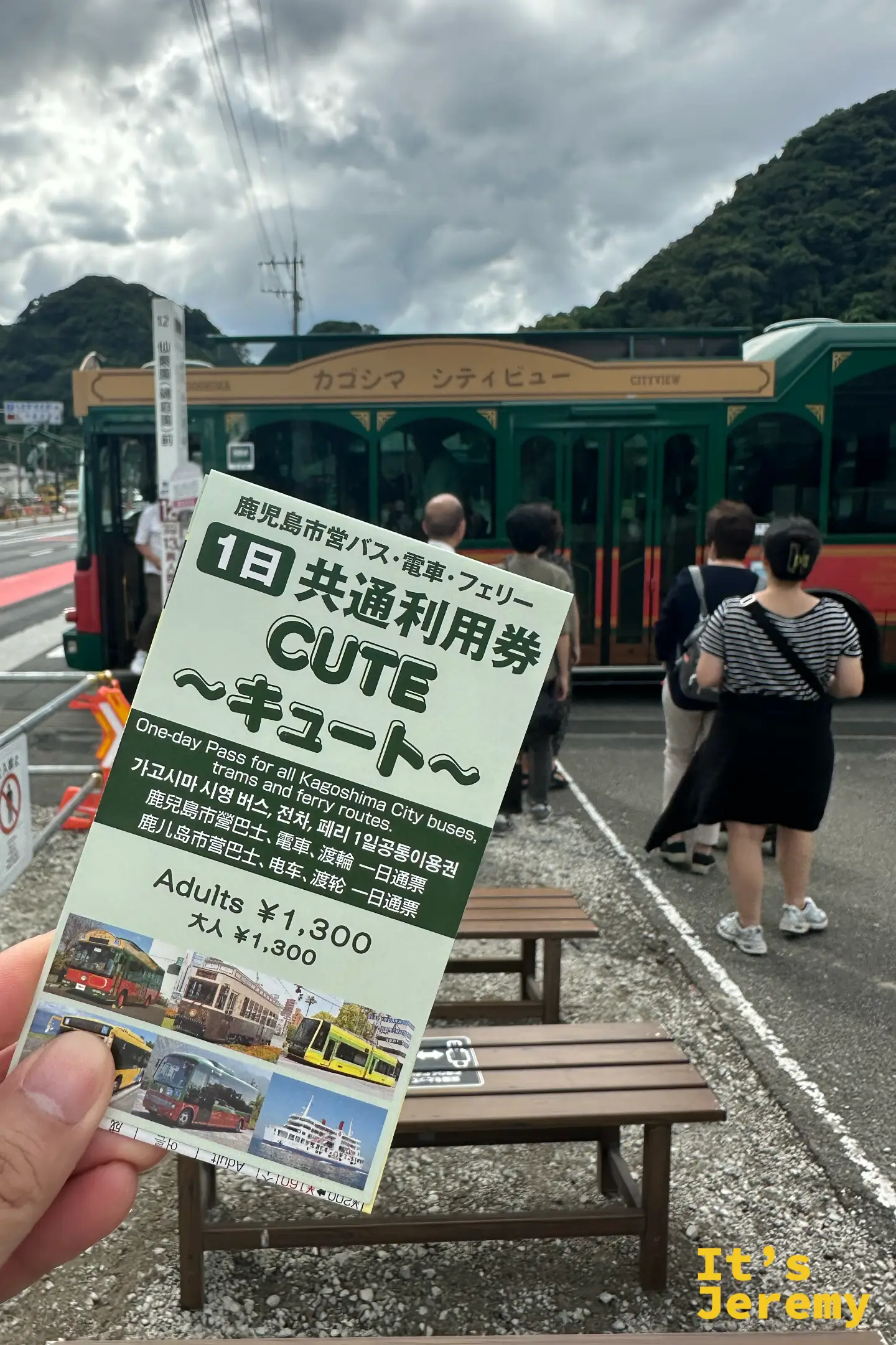 手持鹿兒島「CUTE」一日共通利用券，券面標示可搭市營巴士、電車與渡輪，成人 1,300 日圓，背景是綠紅配色的觀光巴士