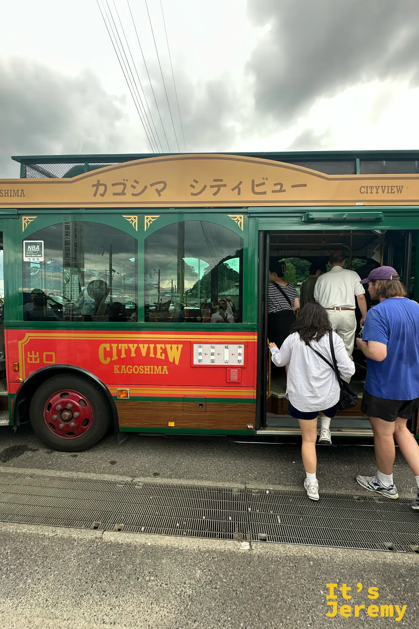 綠紅配色的「カゴシマ シティビュー」觀光巴士停在站牌旁，多名遊客正排隊上車，車身標著中央駅、城山、仙巌園的循環路線