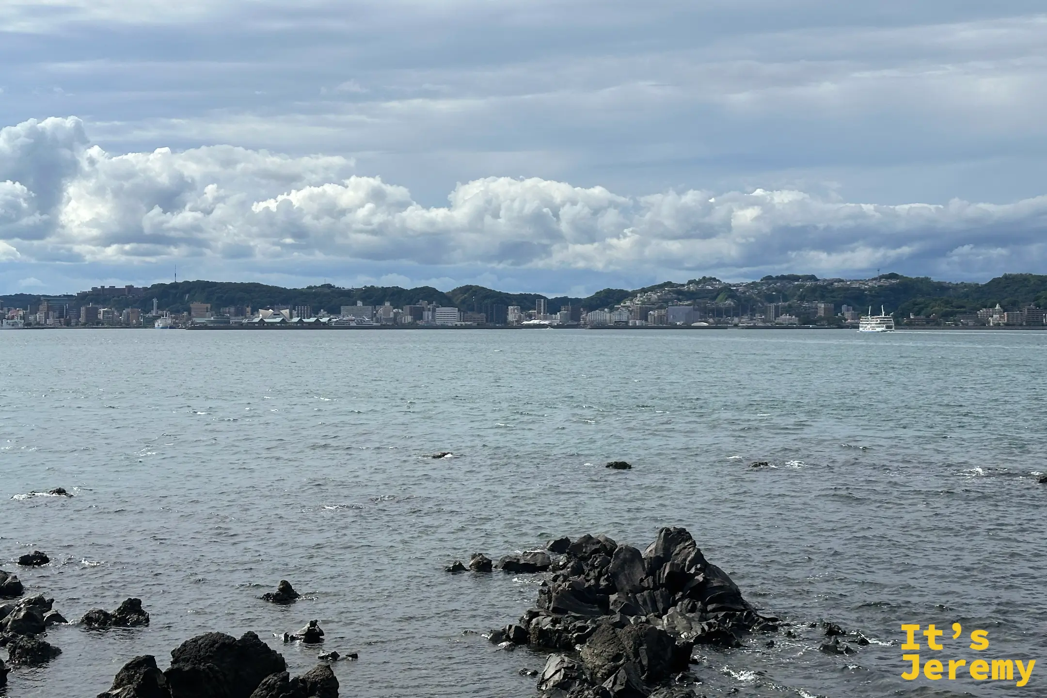 從溶岩なぎさ遊歩道望向錦江灣對岸的鹿兒島市區，前景是黑色熔岩礁岩