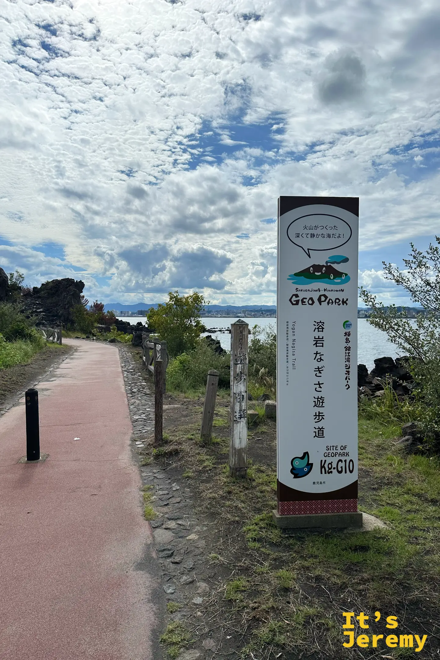 溶岩なぎさ遊歩道入口的地質公園立牌，寫著「火山造出來的、又深又靜的海」，紅色步道沿著黑色熔岩海岸延伸，對岸是鹿兒島市天際線