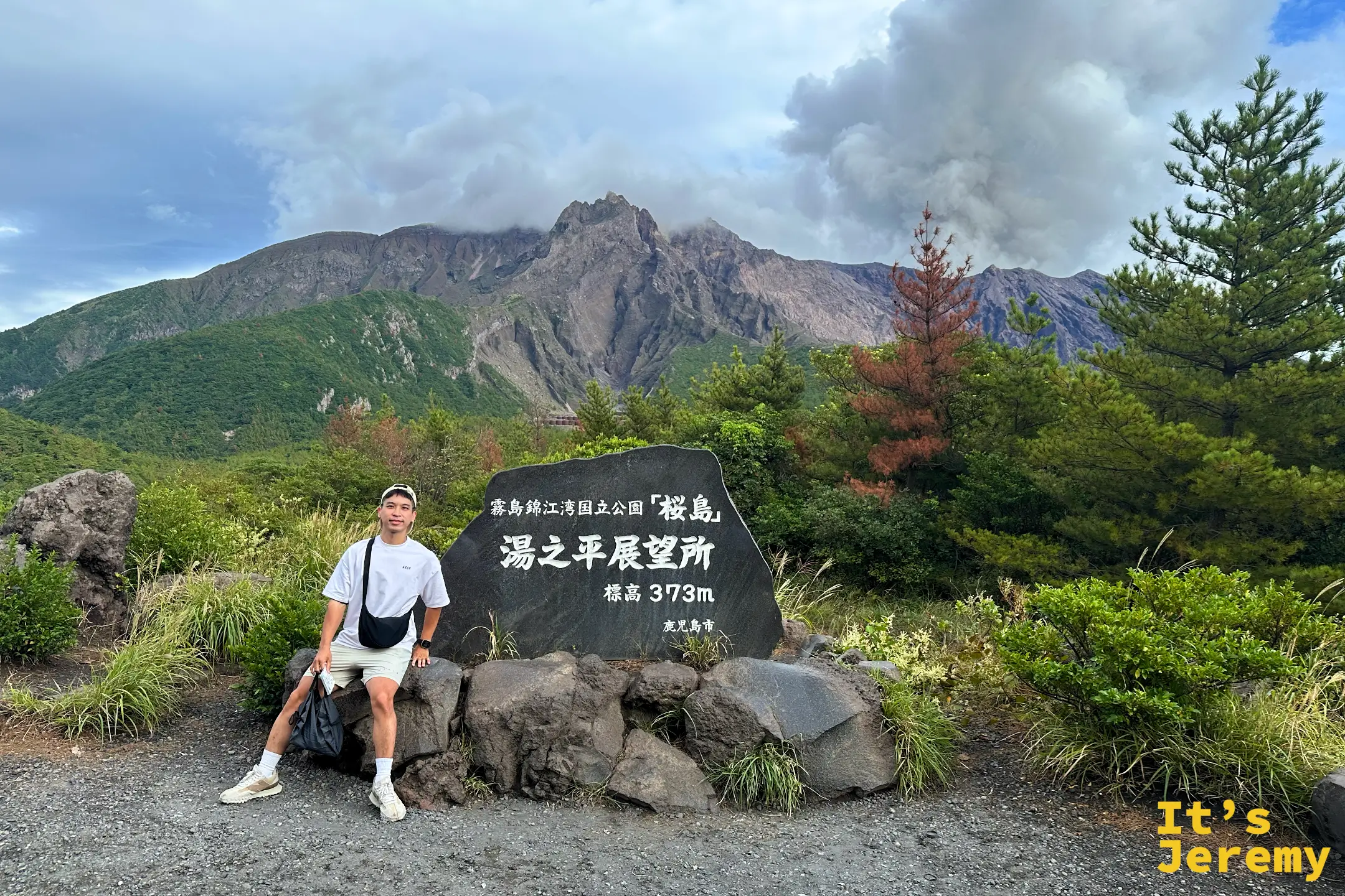 Jeremy 坐在臨海草地的長椅上，戴著墨鏡與帽子微笑，身後隔著平靜的錦江灣就是雲帽下的櫻島火山