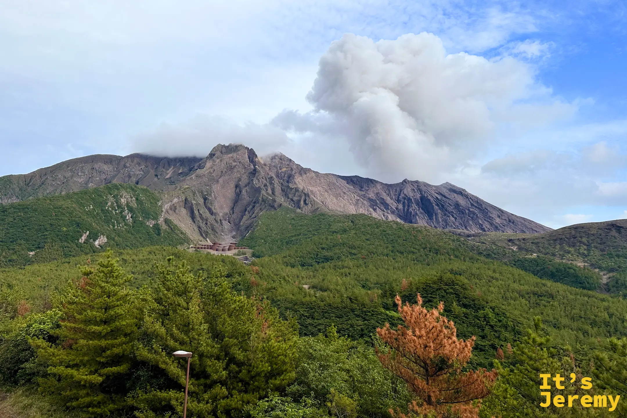 從櫻島西側望向錦江灣，前景是黑色熔岩與草地構成的荒地，海面上有一座小小的燈塔島，對岸是鹿兒島市區