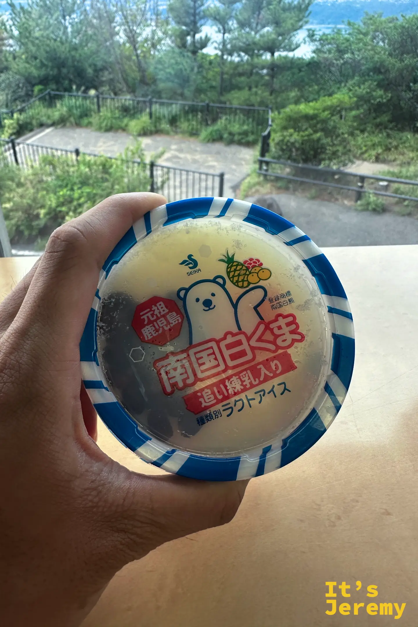 Jeremy 坐在「櫻島 湯之平展望所 標高373m」的石碑旁，身後櫻島火山高聳、頂端噴著灰煙