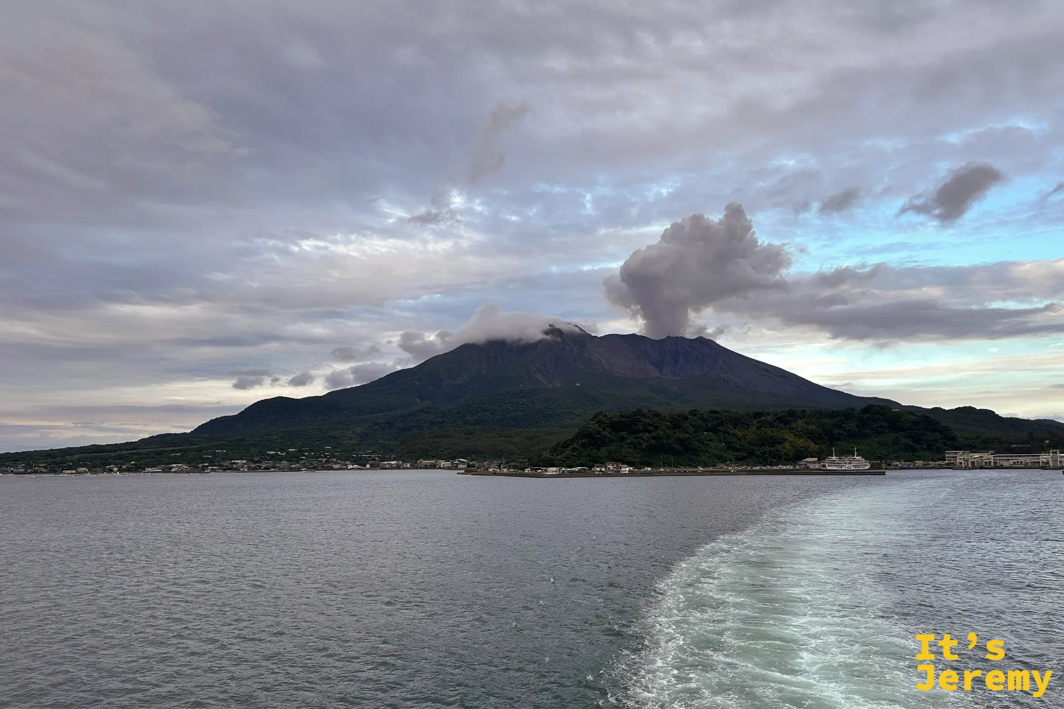 從海上望向櫻島，山頂噴出大片灰色火山煙，天空是清晨的藍灰色調
