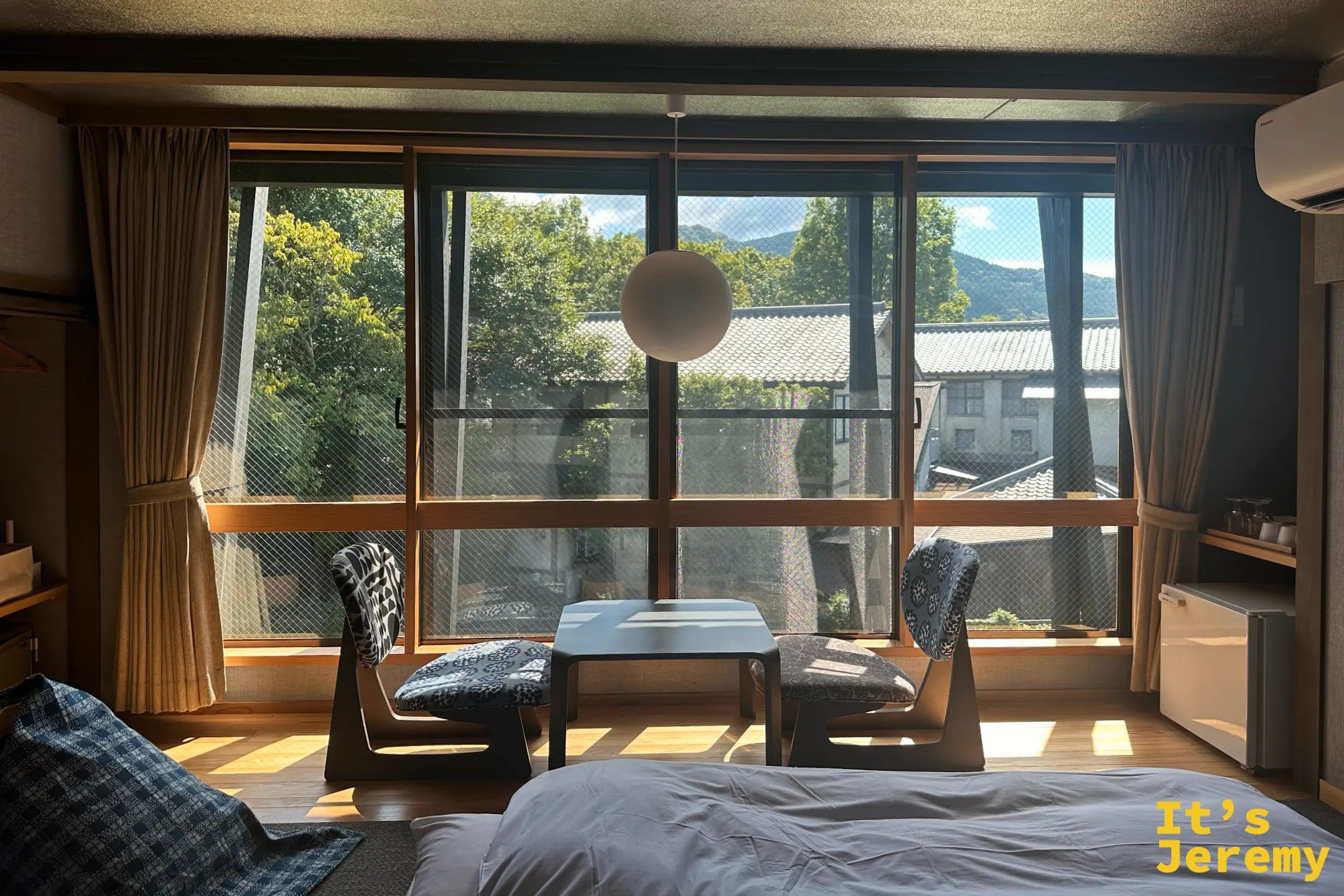 榎屋旅館房間窗邊，兩張矮腳椅與茶几對著落地窗，窗外是綠樹、瓦頂民宿與遠山