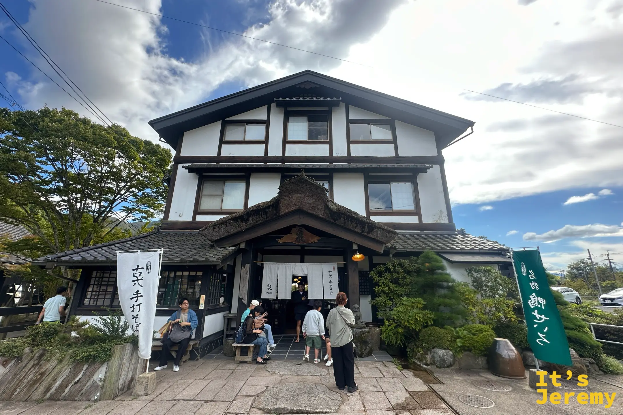 泉的正面全景，白牆黑木三層老屋，門口白暖簾寫著泉字，兩側旗幟與等候的客人