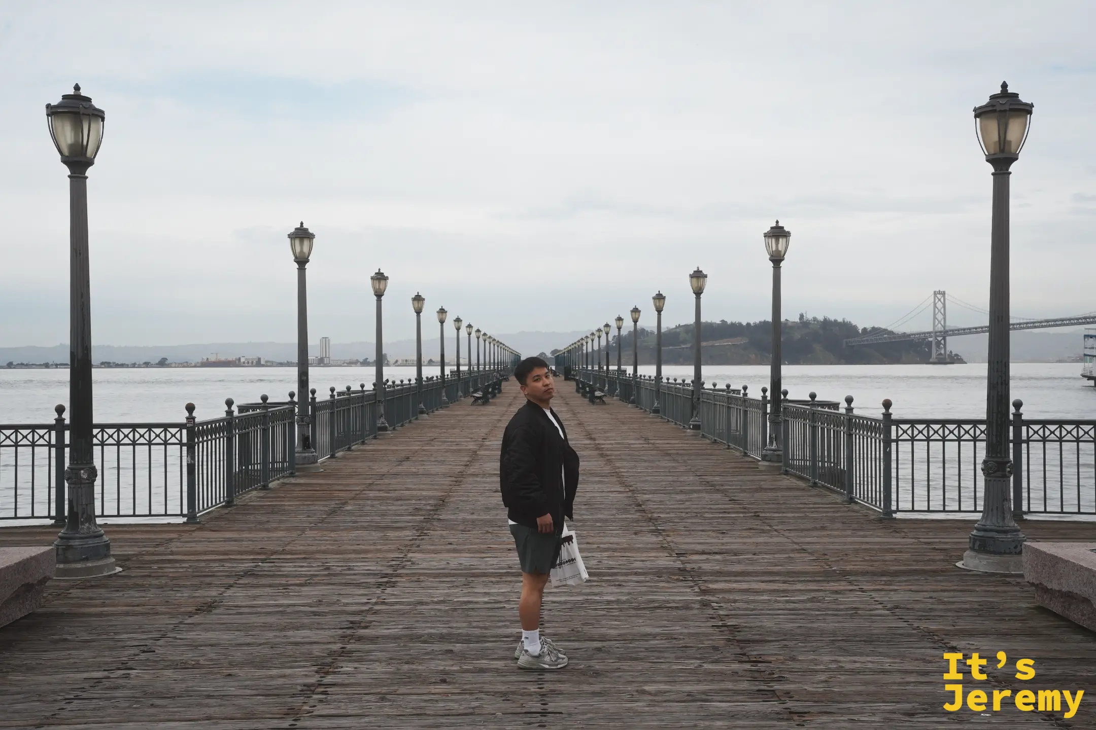 Jeremy 站在 Pier 7 的木棧步道上，兩排老式路燈延伸向海灣，遠方是海灣大橋