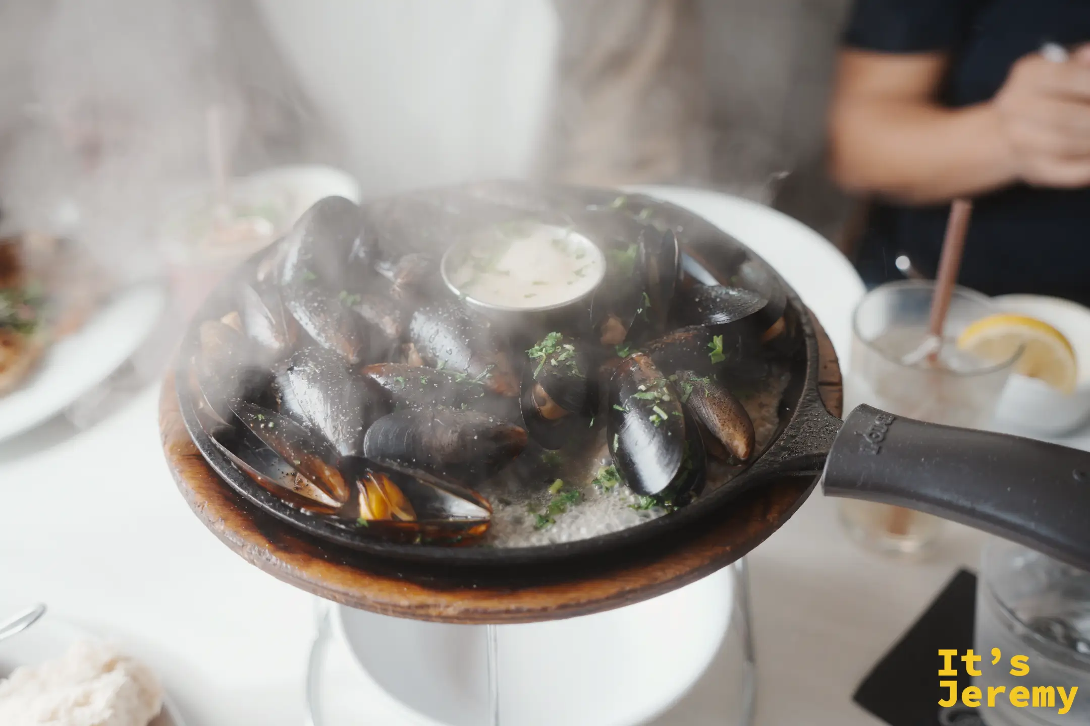 一鍋冒著白煙的蒜香奶油蒸淡菜，黑殼淡菜排滿鑄鐵鍋，中央附一小杯醬汁
