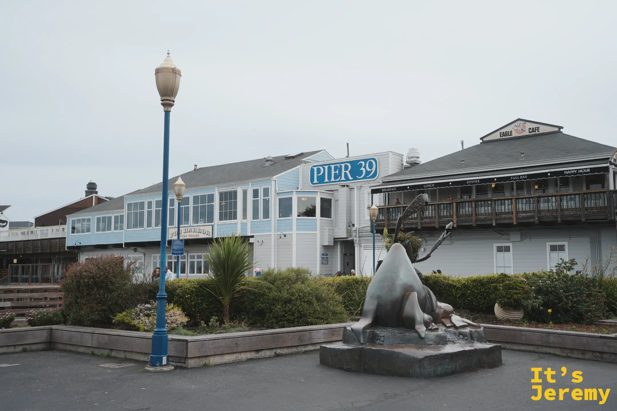 PIER 39 招牌下的廣場，前方有一座青銅海獅雕塑，右側是 Eagle Cafe，左側是 Fog Harbor Fish House