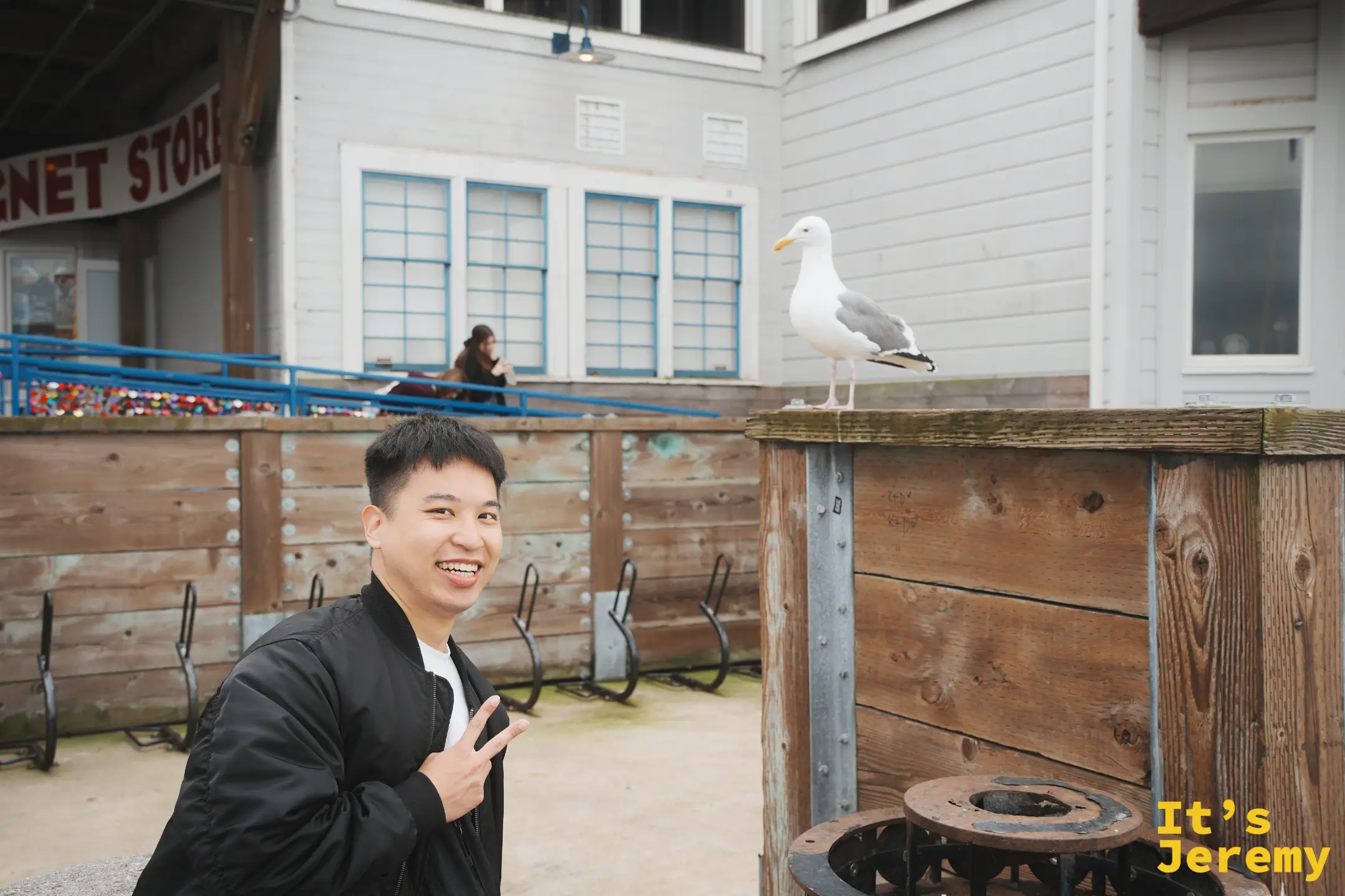 Jeremy 穿黑色飛行外套比出手勢微笑，右邊木樁上站著一隻不怕人的大海鷗