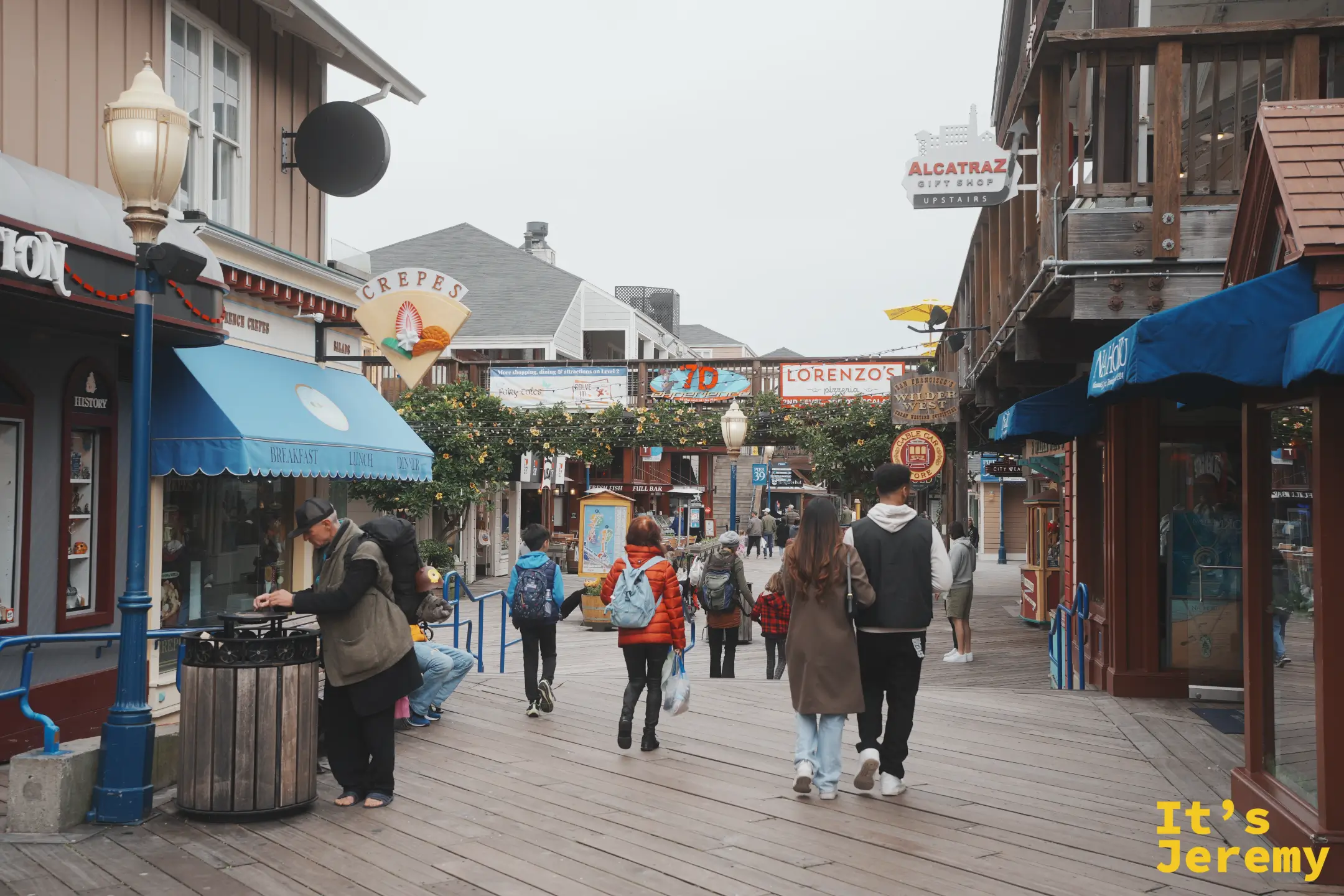 PIER 39 街道層的店家，左側是藍色遮陽棚的可麗餅店，遠處掛著 Alcatraz 禮品店與 Lorenzo