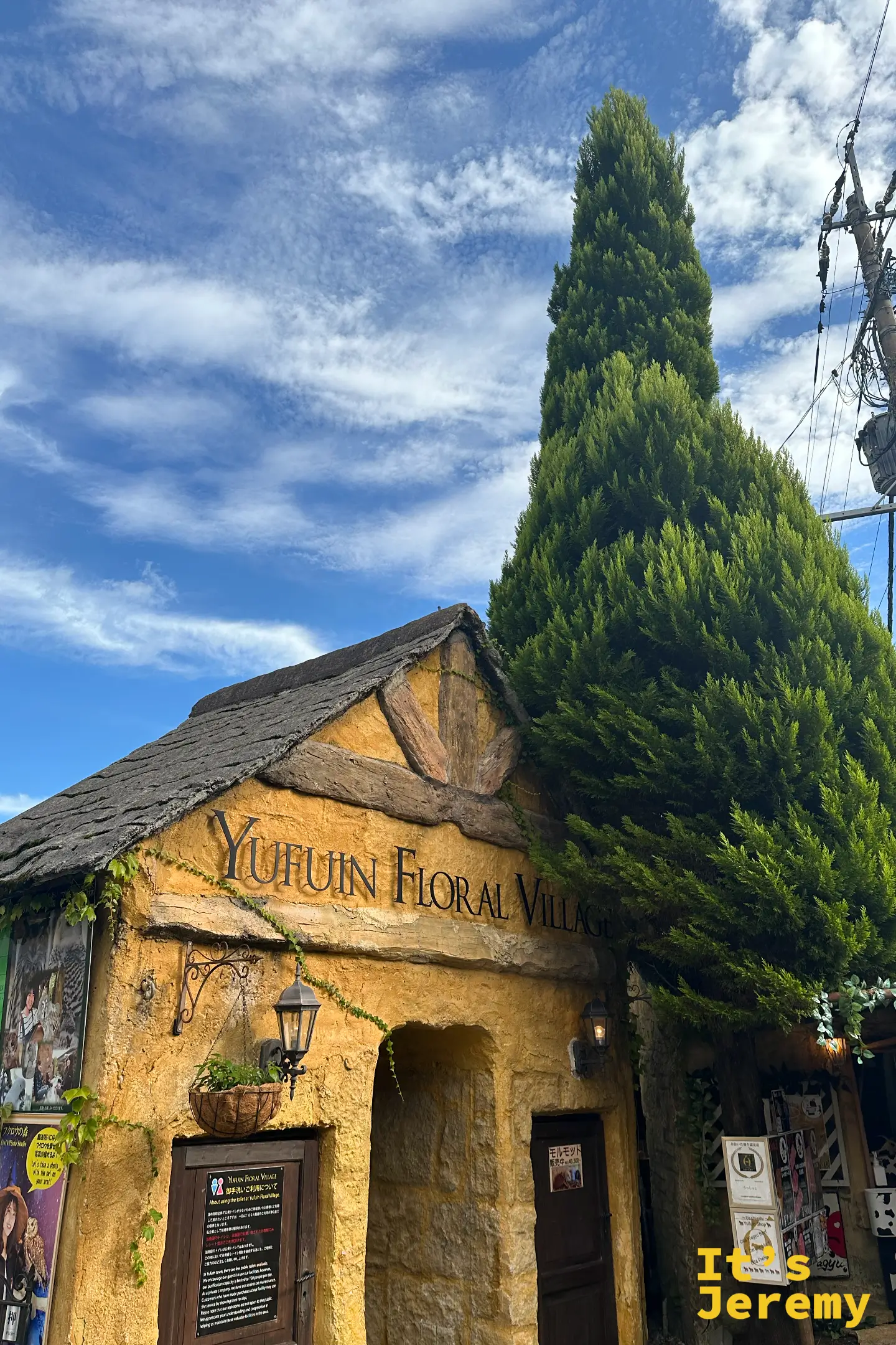 Yufuin Floral Village 入口招牌特寫，黃牆上有掛燈與吊籃植栽