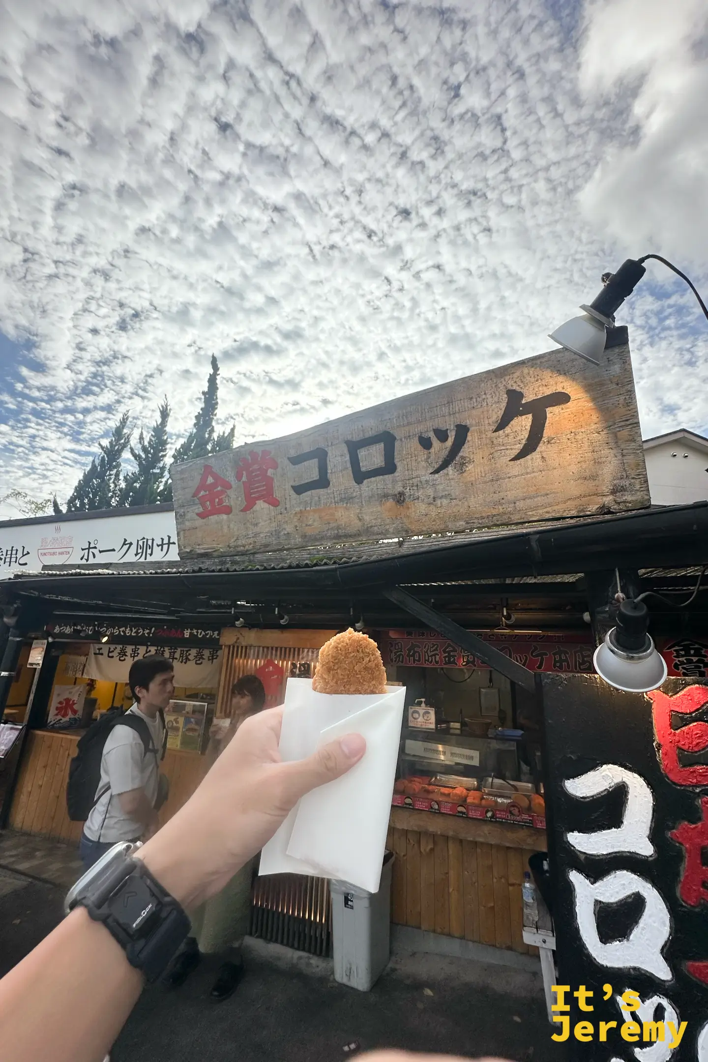 手拿金賞可樂餅，背景是掛著木招牌的金賞コロッケ店面