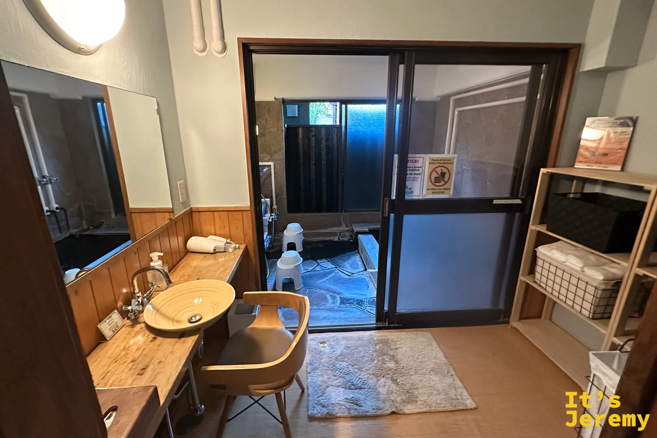 湯屋外的梳洗區，木質檯面搭配陶製洗手盆與設計椅，透過玻璃拉門連著石造浴池