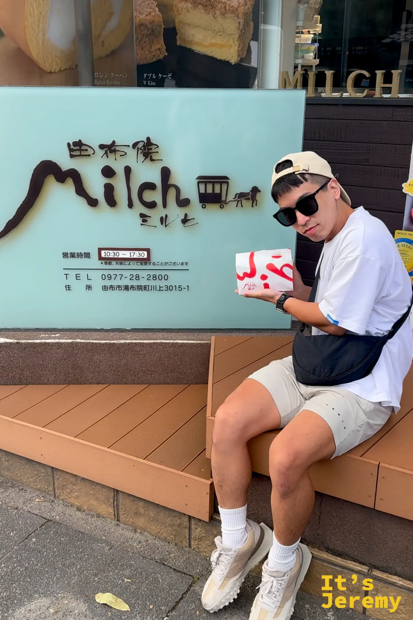 Jeremy 坐在由布院 Milch 招牌前，手拿一袋 Milch 的外帶商品